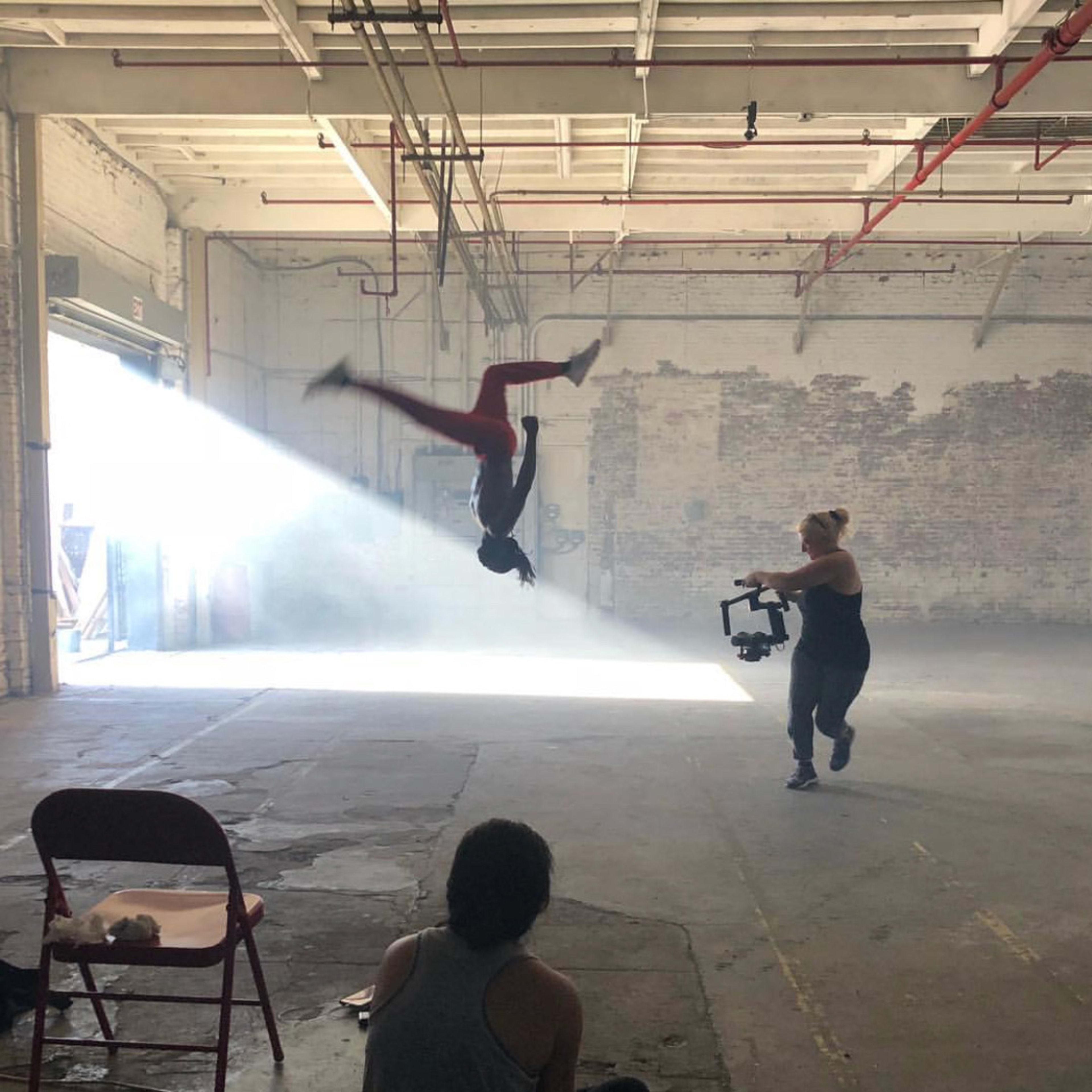 A person performs an acrobatic flip in an industrial space while another individual captures the moment with a camera.