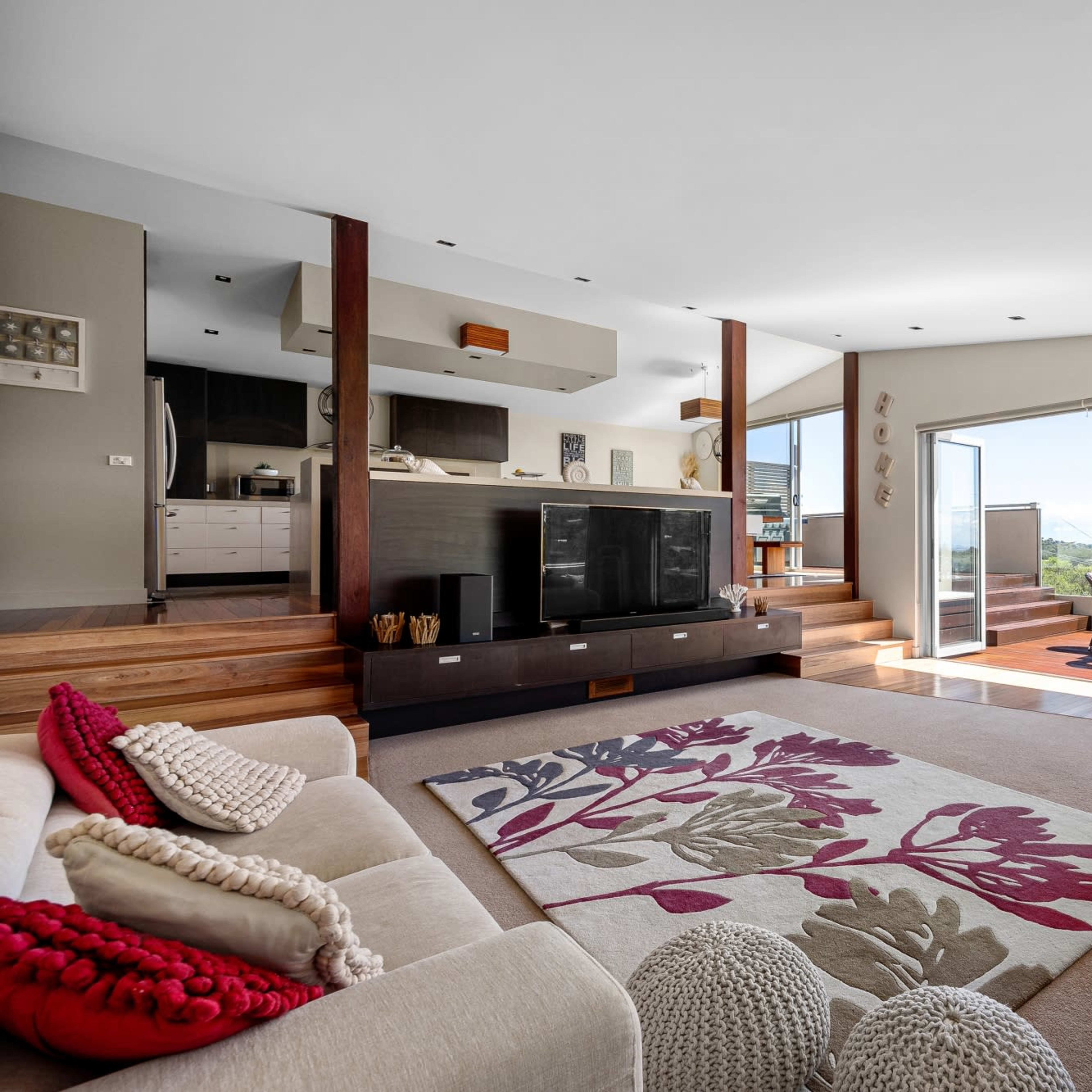 The living room features a spacious layout with a large area rug, two sofas, a modern kitchen in the background, and large sliding glass doors that open to a view outside.