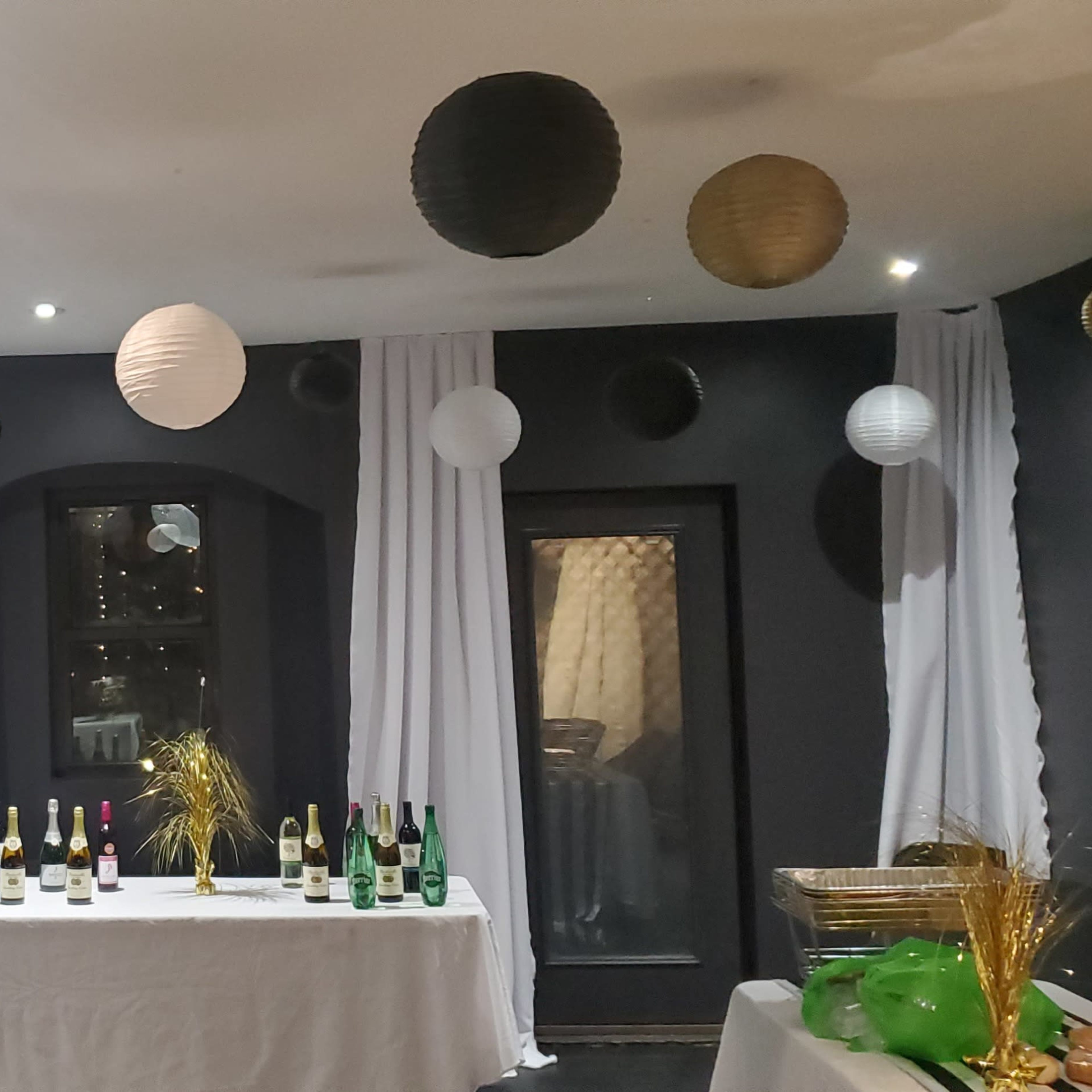 The room features white and black paper lanterns hanging from the ceiling, with tables covered in white linens displaying bottles of wine and champagne.