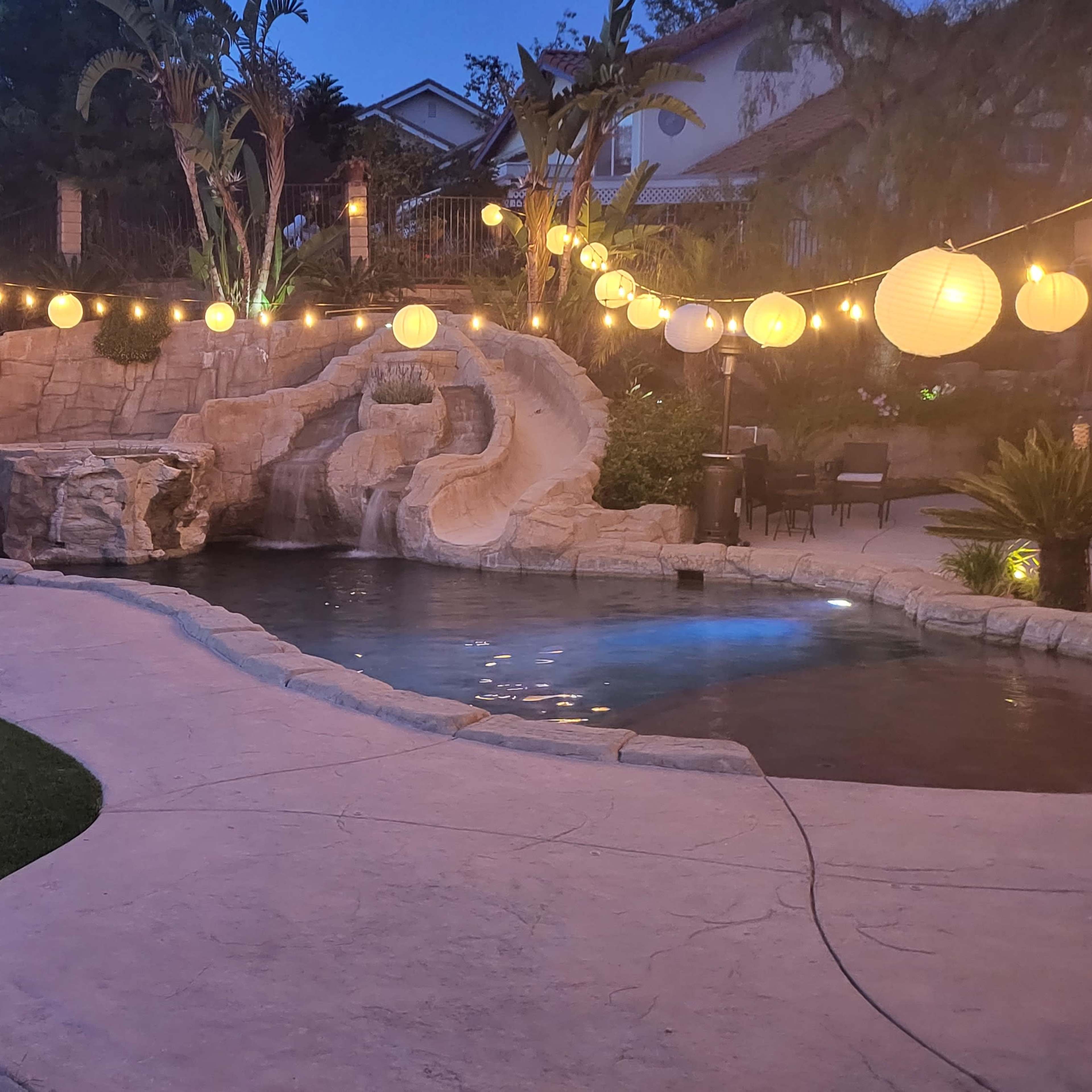 A backyard pool area with a rock formation, illuminated lanterns strung above, and a well-maintained lawn.