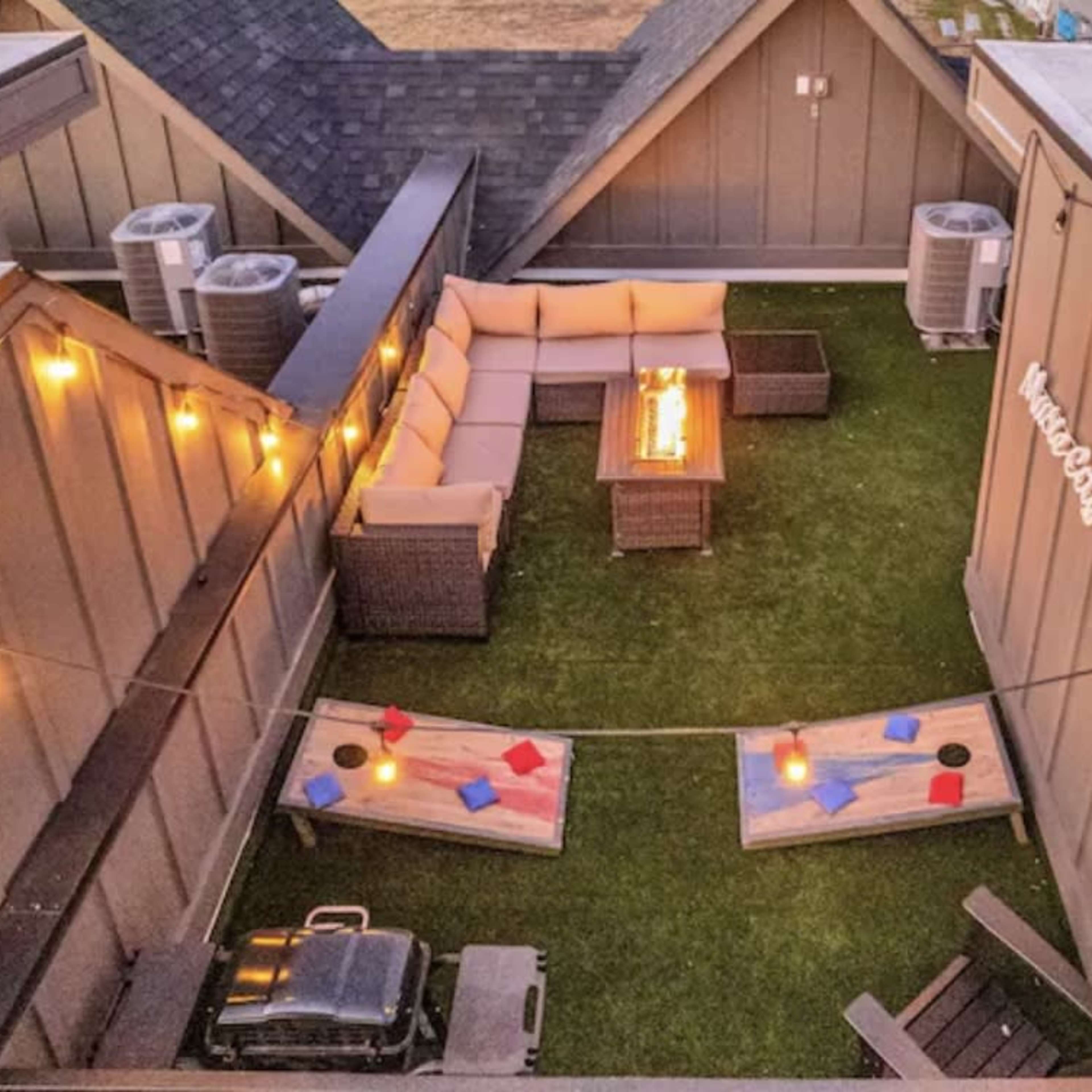 A rooftop patio with a seating area, string lights, and two cornhole boards on a grassy surface.