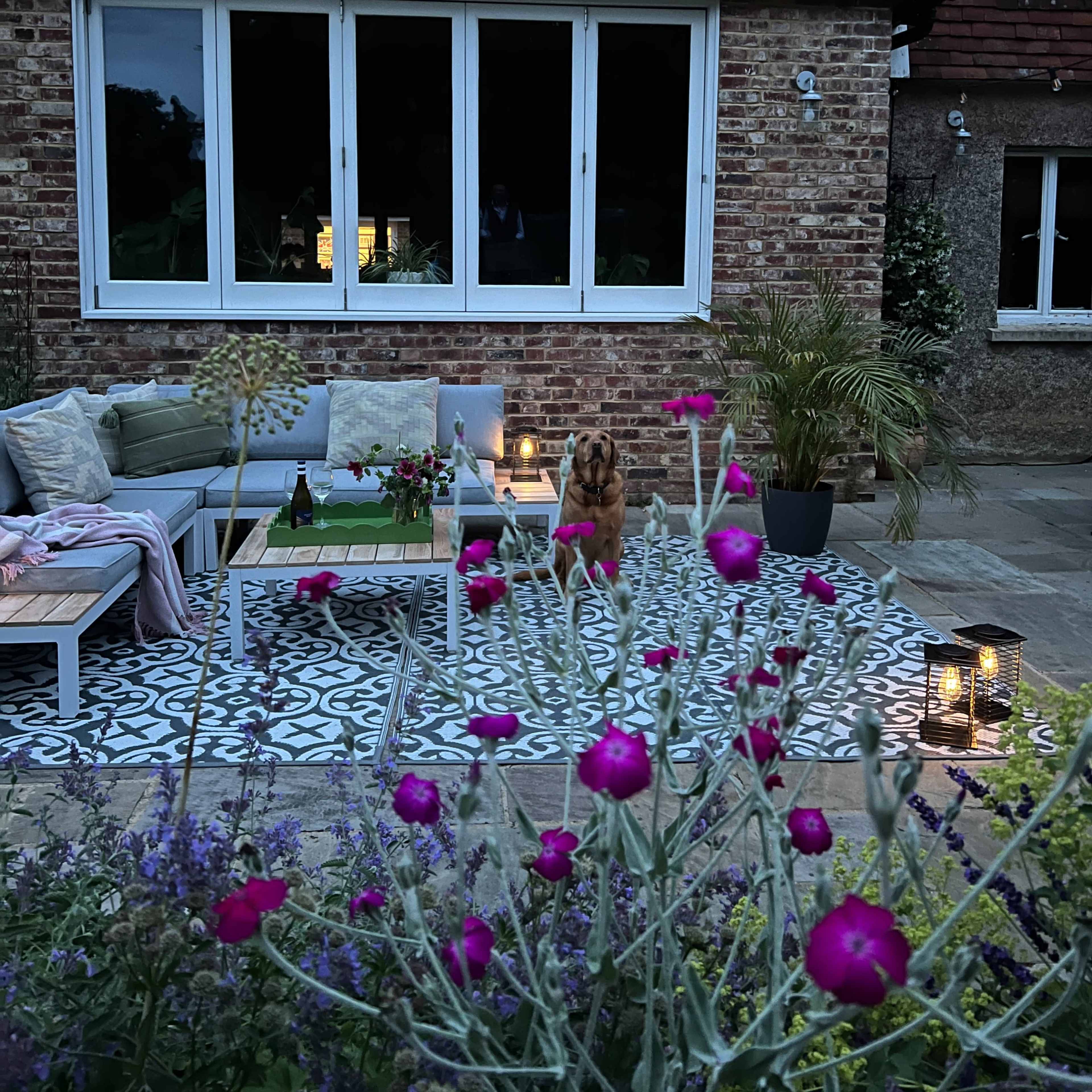 A cozy outdoor seating area with a dog, surrounded by flowers and illuminated by lanterns, is set against a brick house.