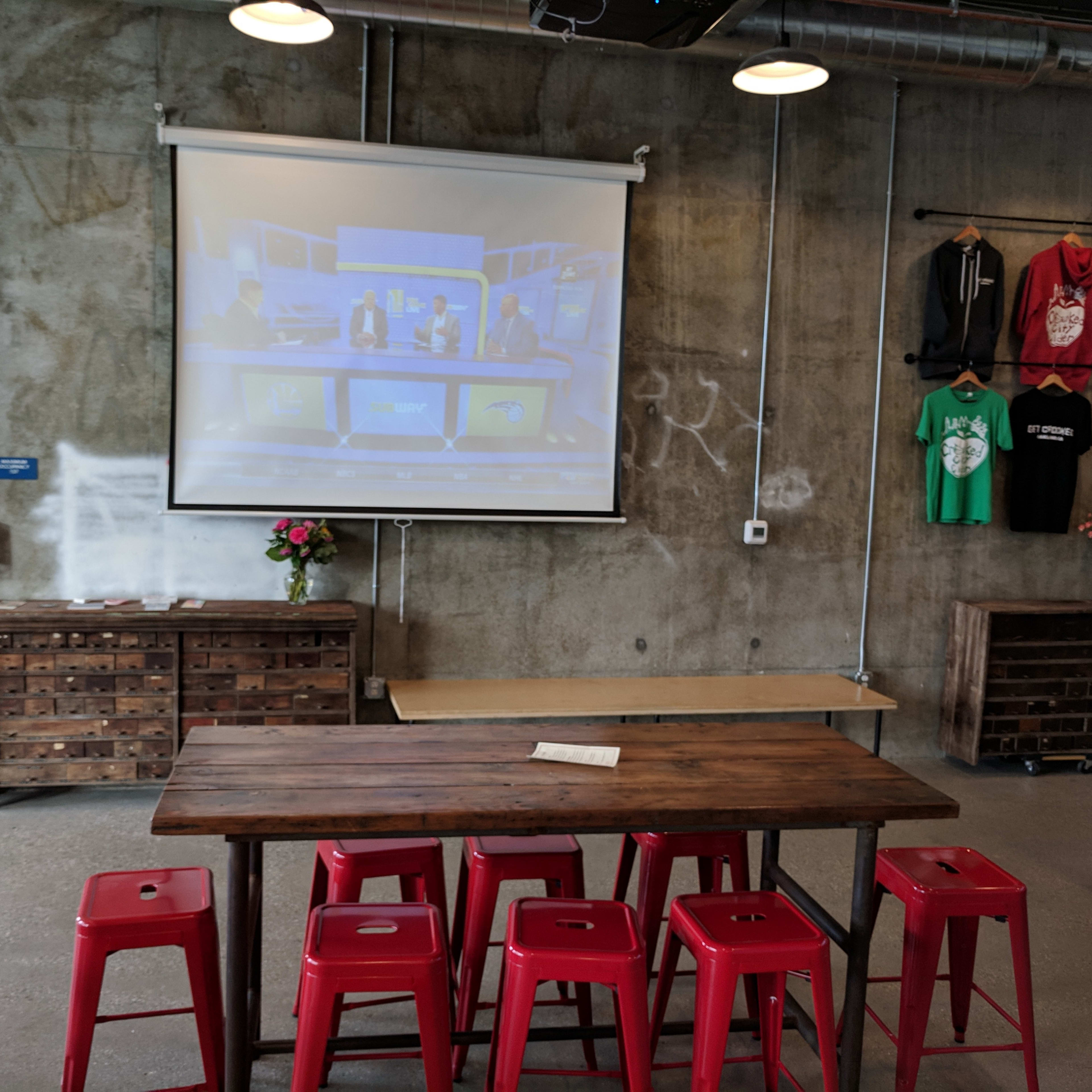 A wooden table with red stools is positioned in front of a projection screen displaying a video in a modern, industrial-style room.