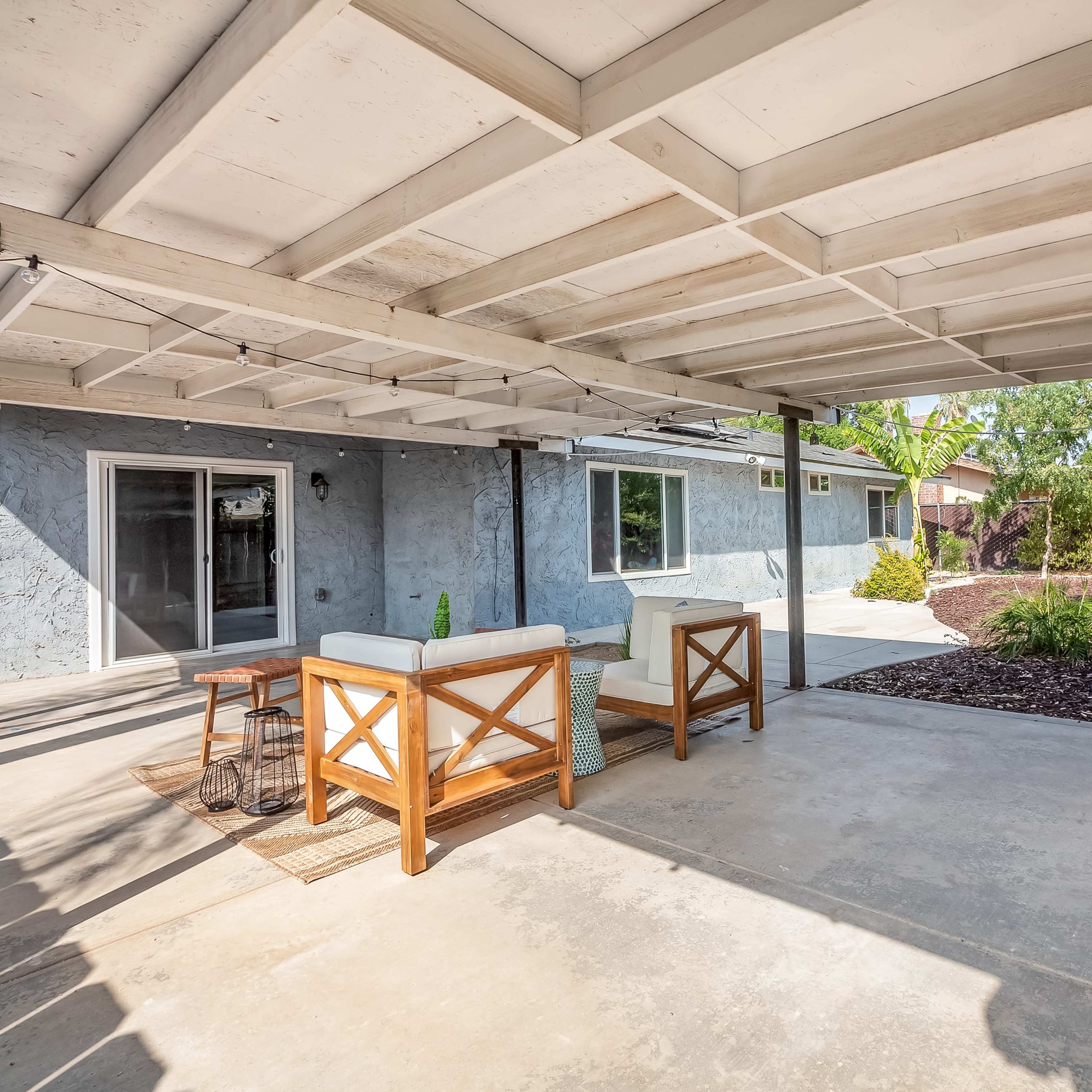 An outdoor patio area with a seating arrangement, adjacent to a residential house surrounded by landscaping.