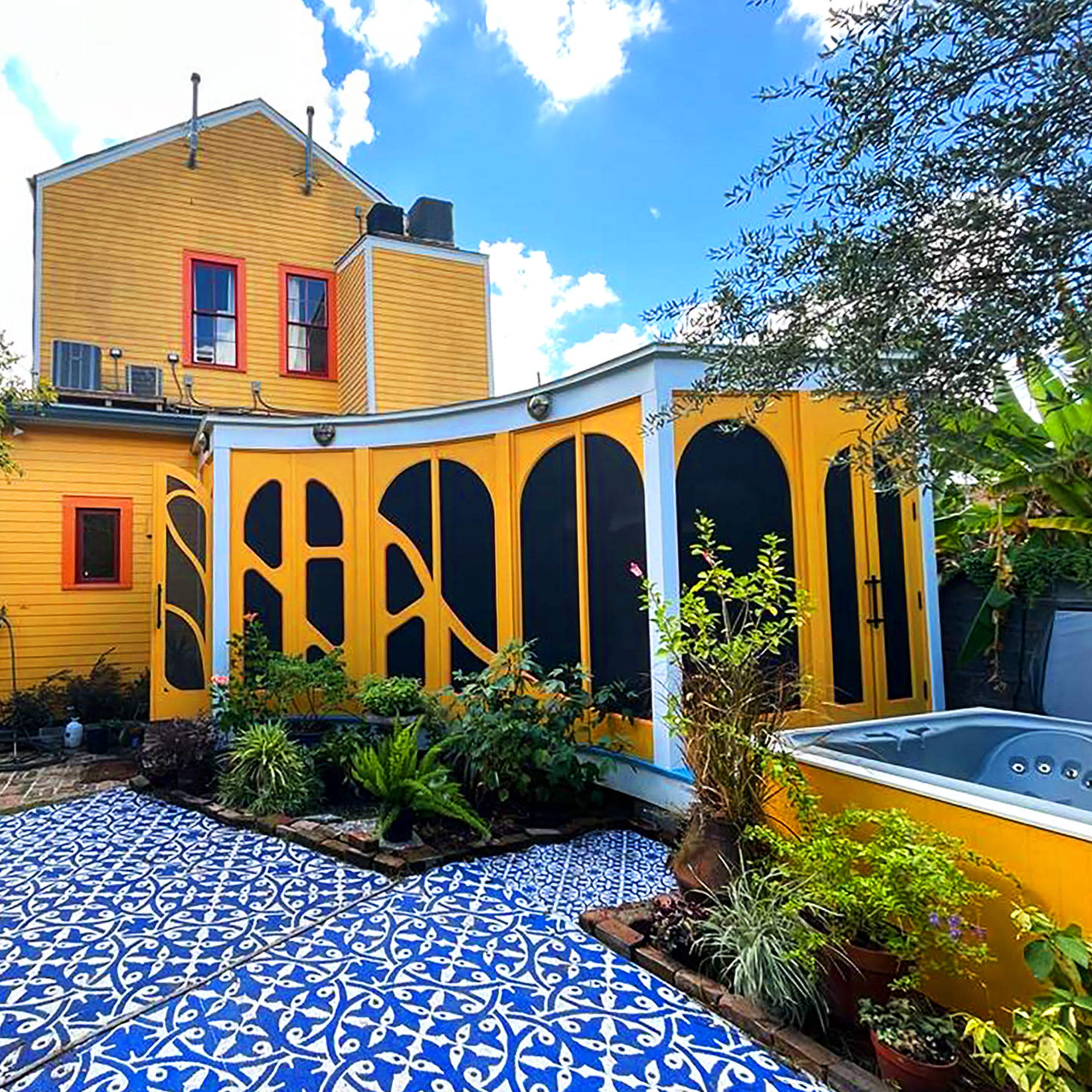 A brightly painted yellow house with red windows features an enclosed patio adorned with intricate blue tiles and surrounded by lush greenery.