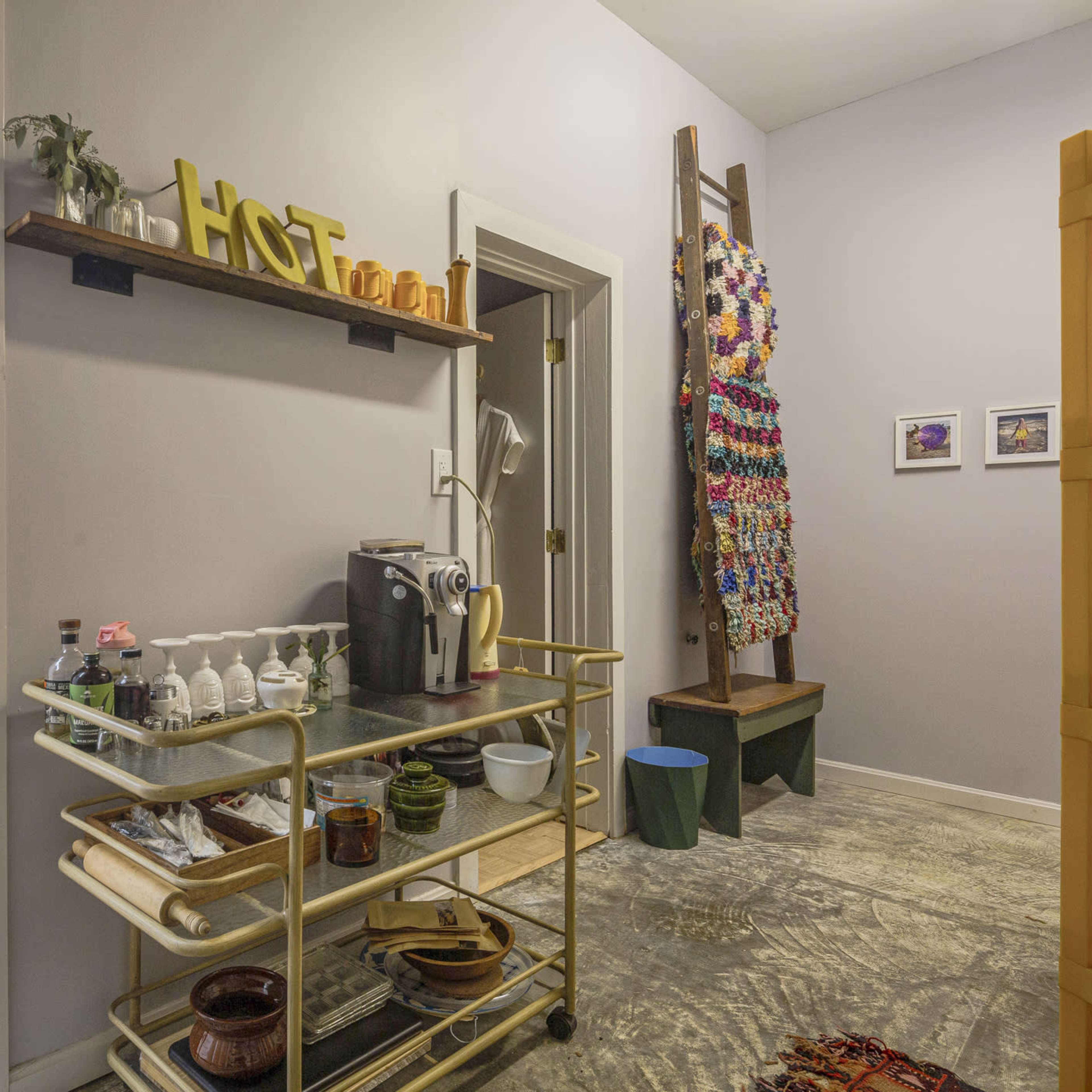 A hallway leading to a room with a kitchen cart displaying various items and a colorful wall hanging made of fabric squares.