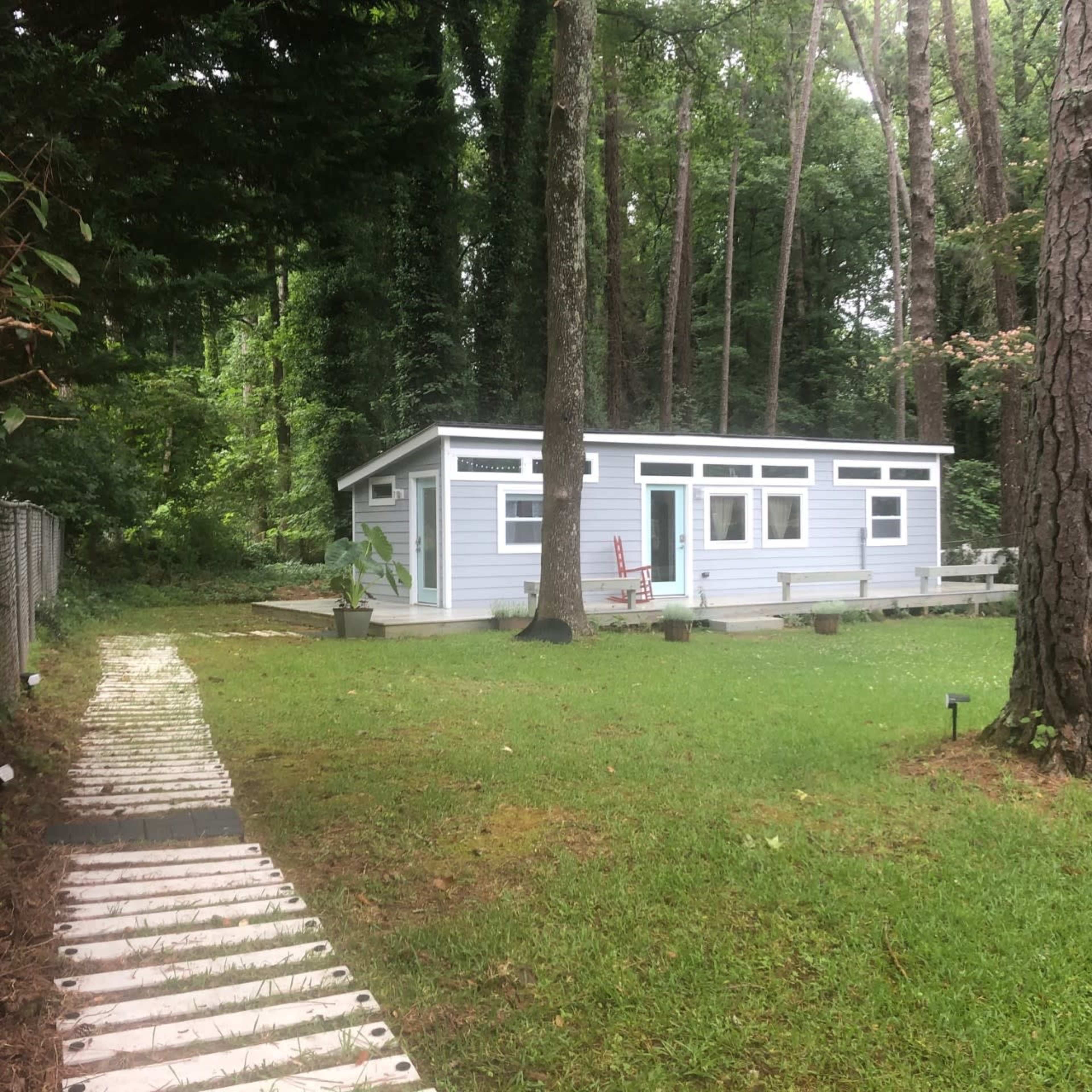 A light blue cabin situated in a grassy area surrounded by tall trees, with a pathway made of wooden planks leading to it.