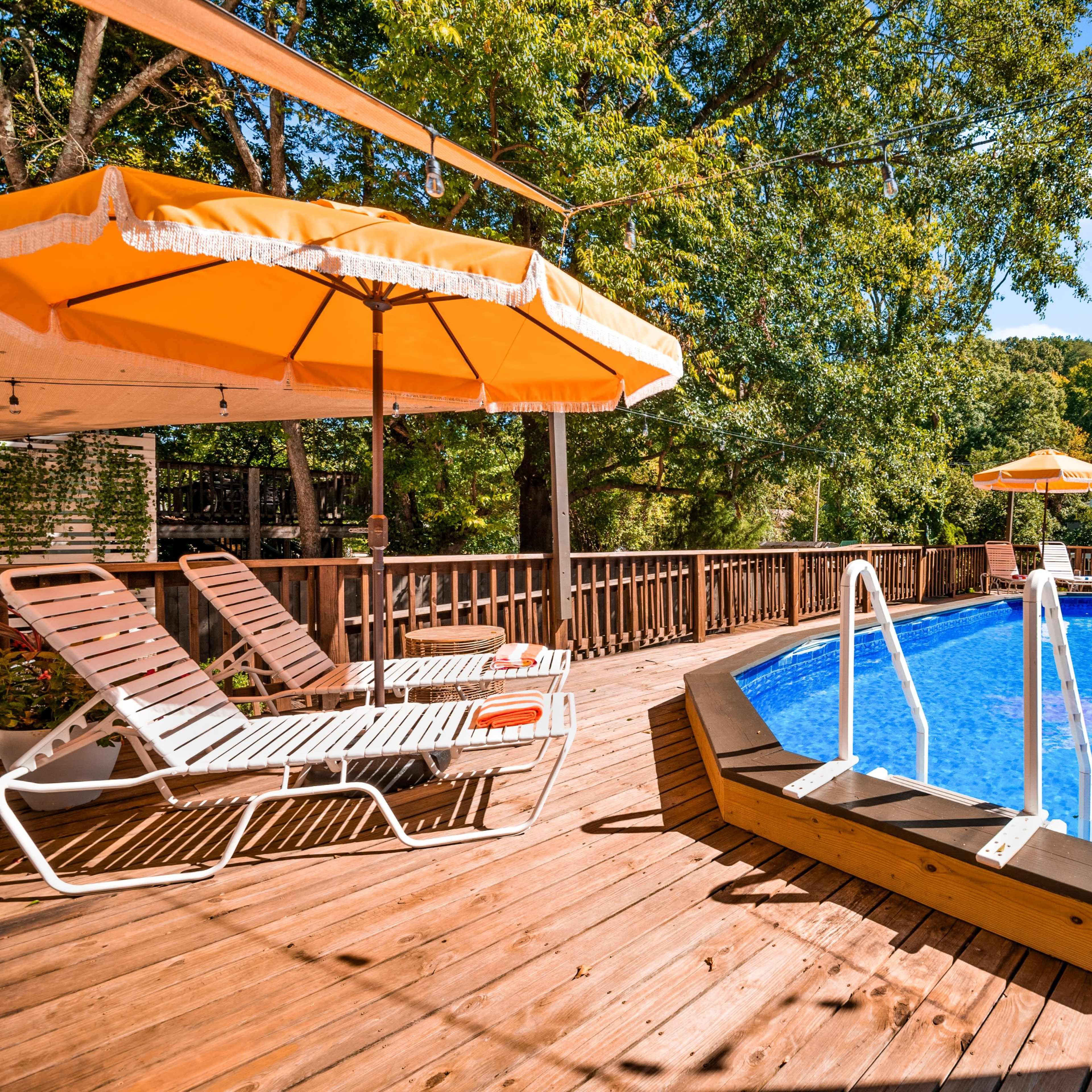 A wooden deck surrounding a circular swimming pool with two orange umbrellas and lounge chairs nearby.