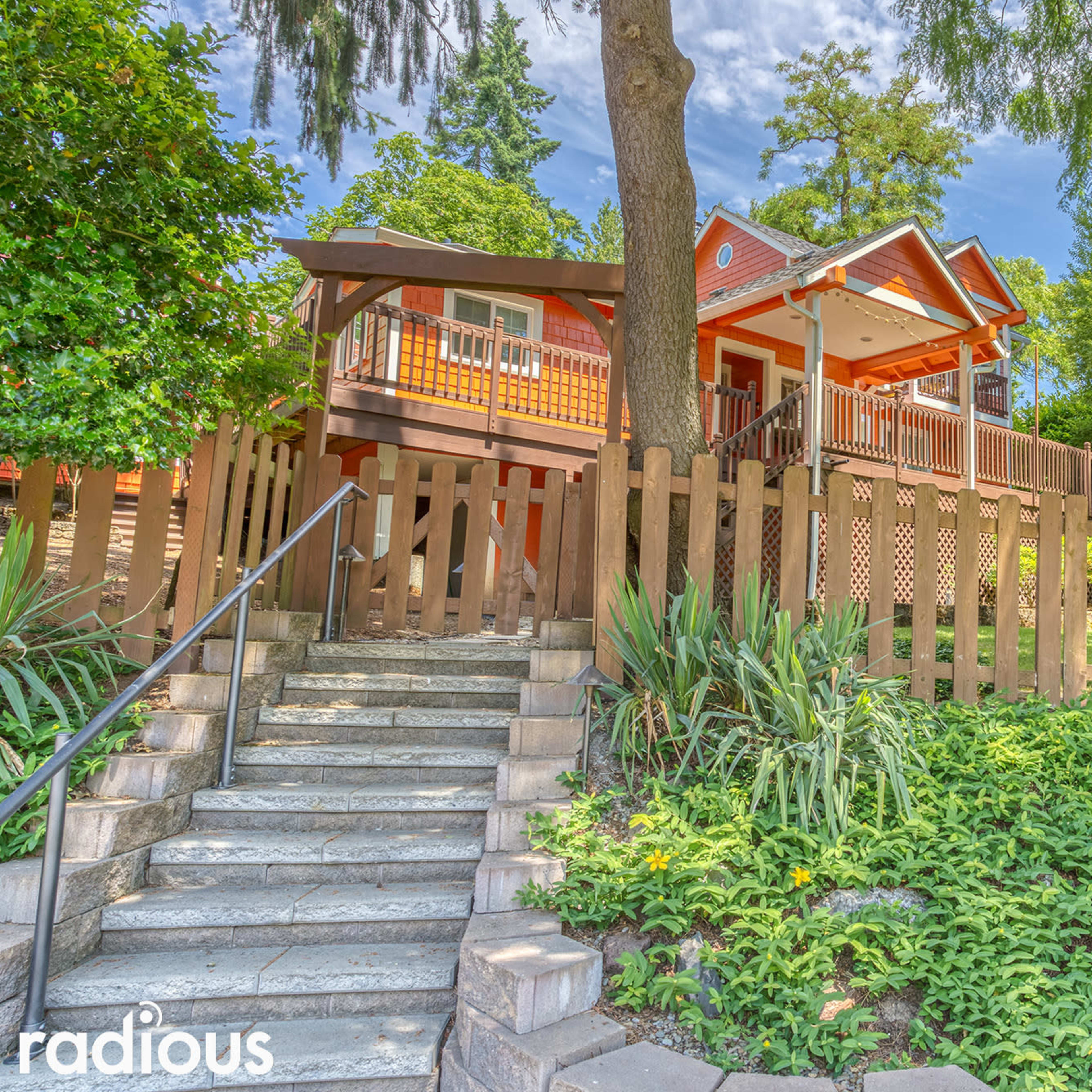 A brightly colored house with a porch and wooden railing is set behind a low fence, surrounded by greenery and trees.