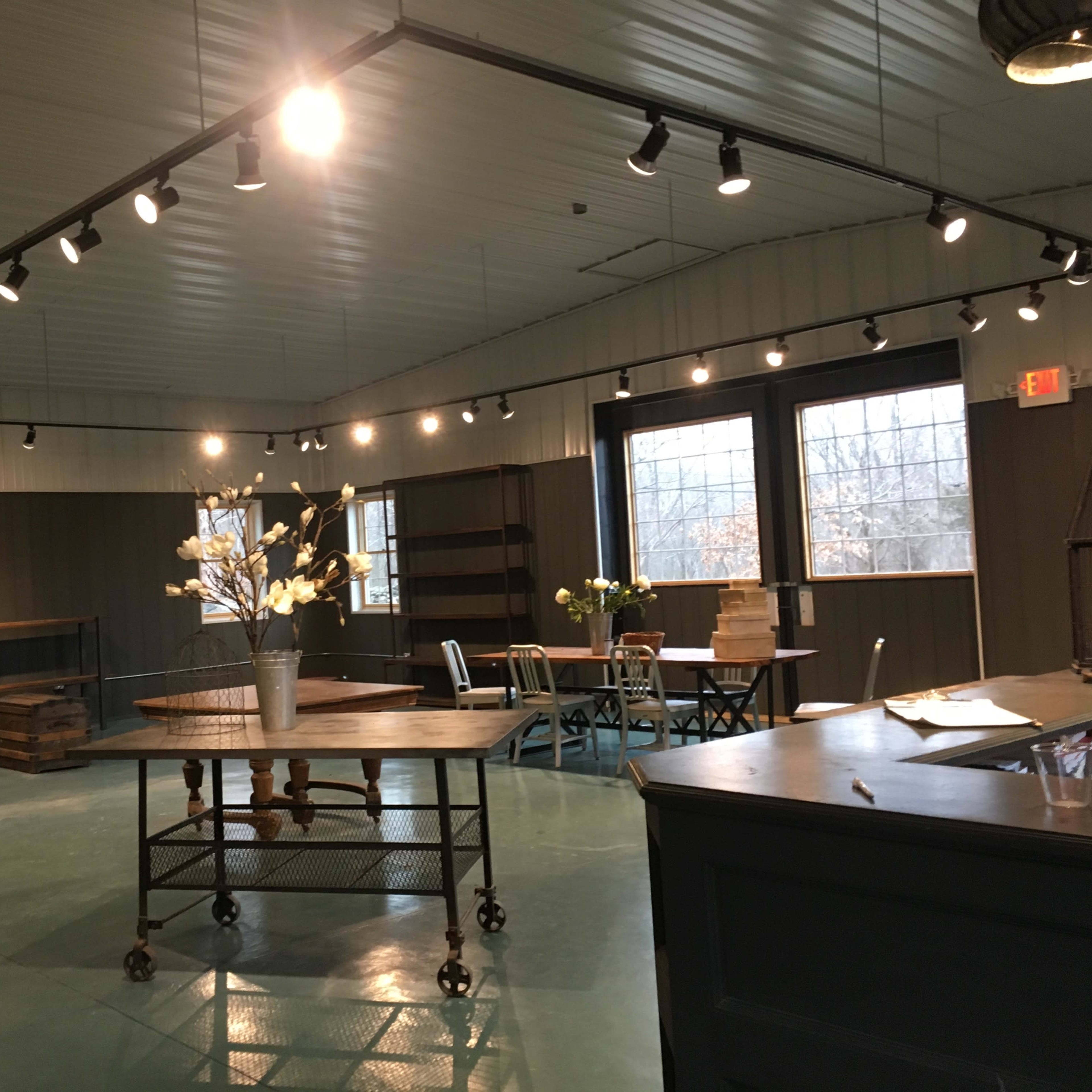 An empty interior space with hardwood floors, featuring several tables, shelves, and large windows illuminating the area.