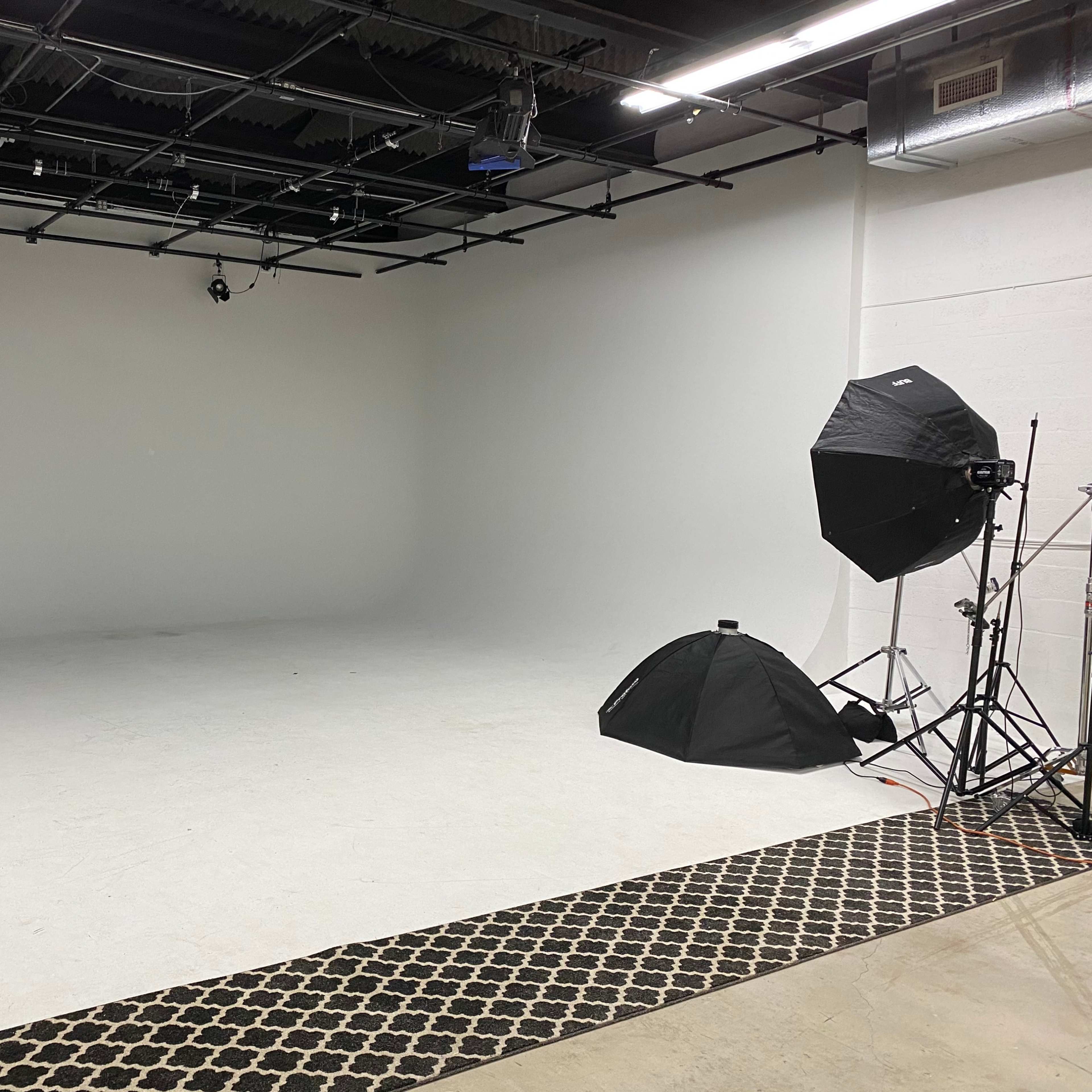 A photography studio features a white backdrop, a black umbrella light setup, and a small tent on the floor.