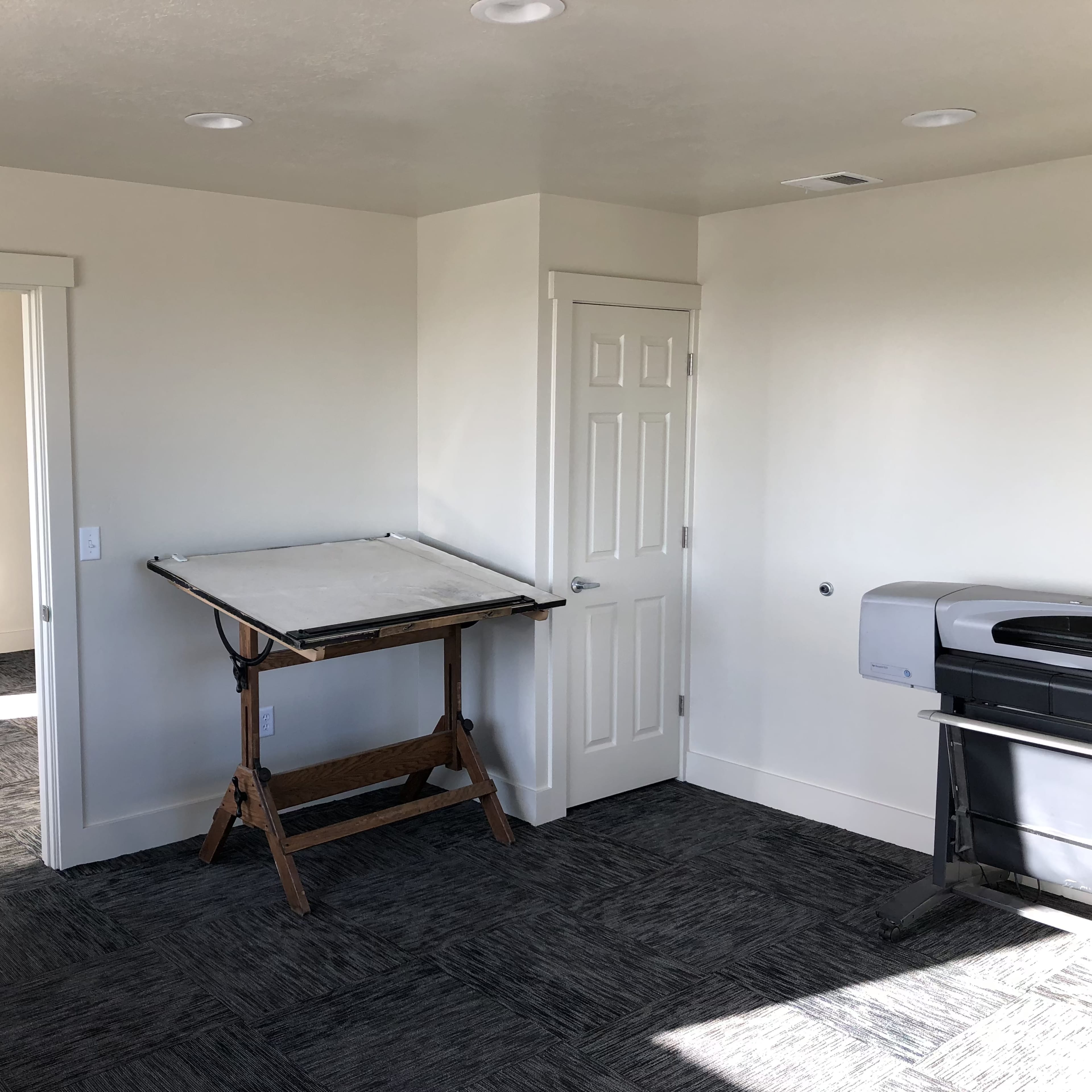 A room with two doors, a drawing board on a stand, and a printer against the wall.