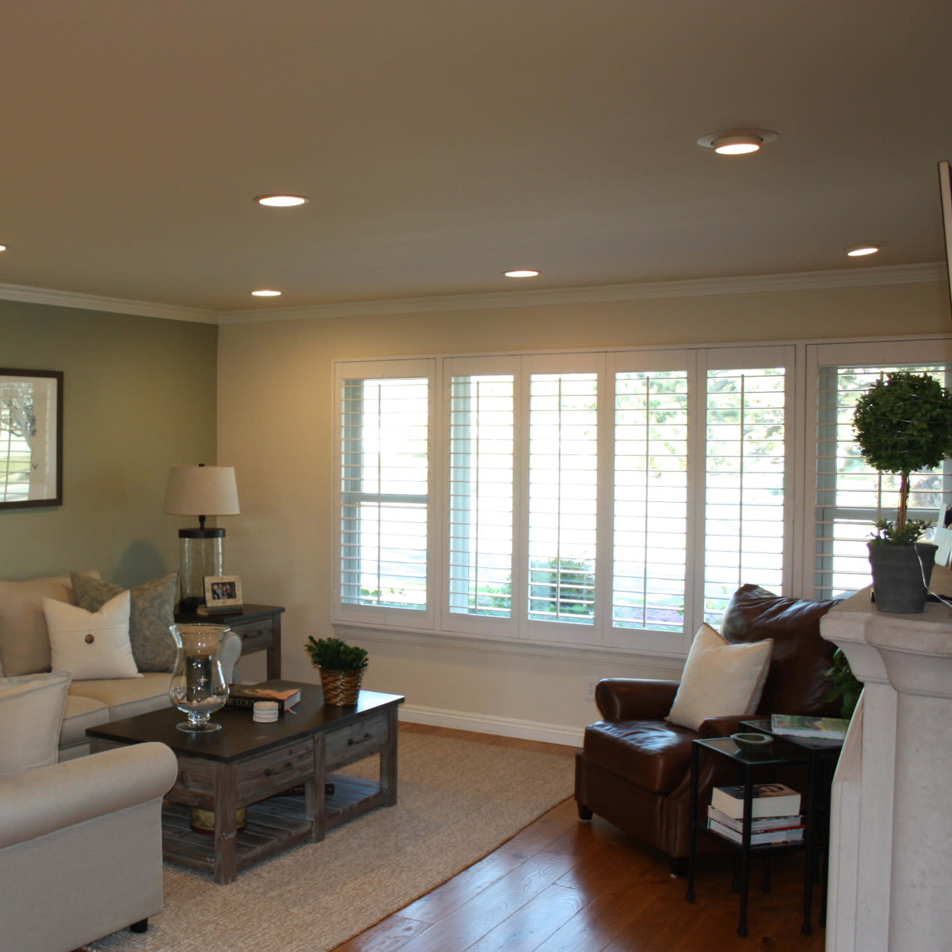 A light-filled living room features a beige sectional sofa, a wooden coffee table, and a chair beside a window with plantation shutters.
