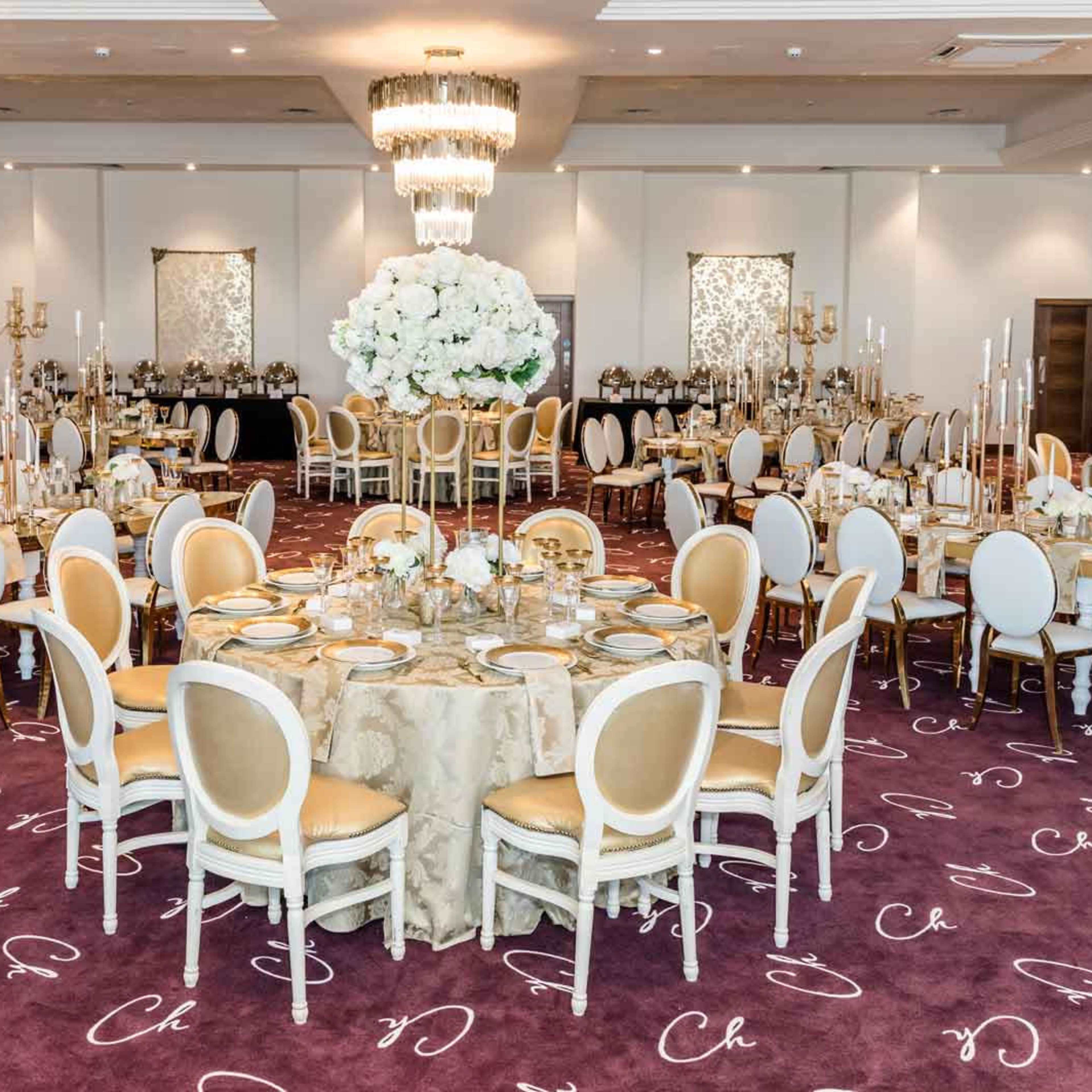 An elegantly set banquet hall featuring round tables with floral centerpieces, surrounded by white chairs on a dark carpet.