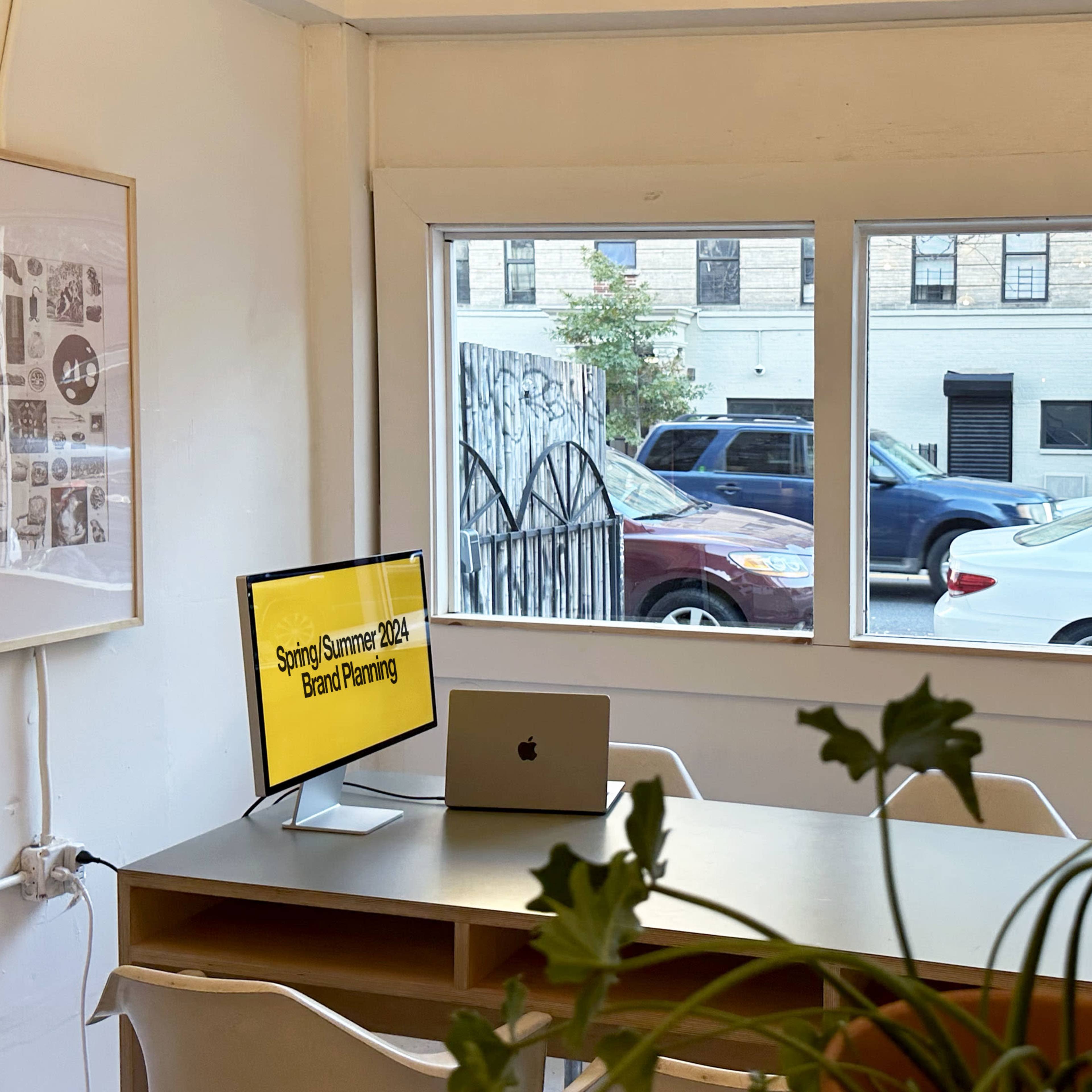 A bright office space with a table and a computer, featuring large windows overlooking a street with parked cars.