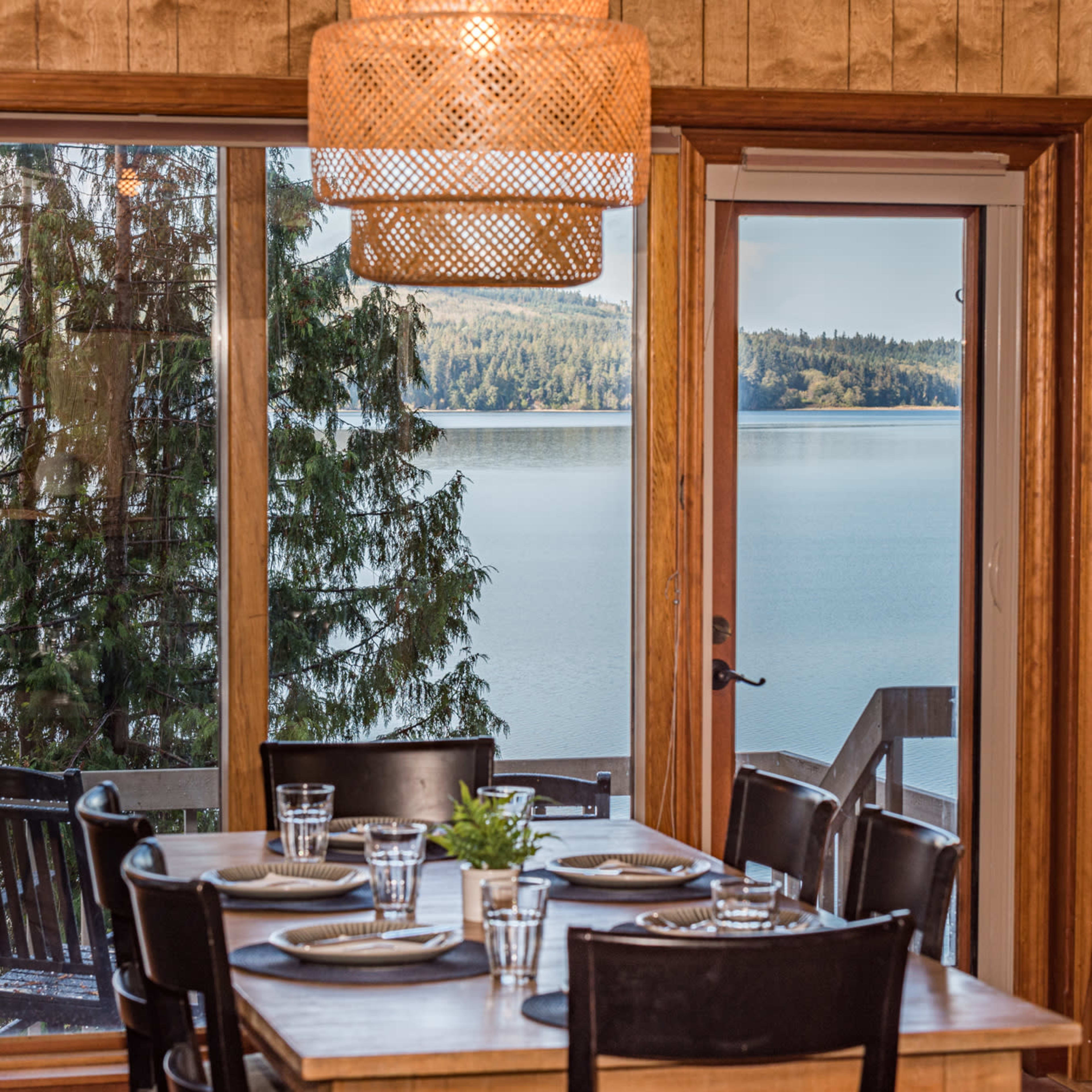 A dining table set with plates and glasses overlooks a serene lake through large windows framed by wooden walls.