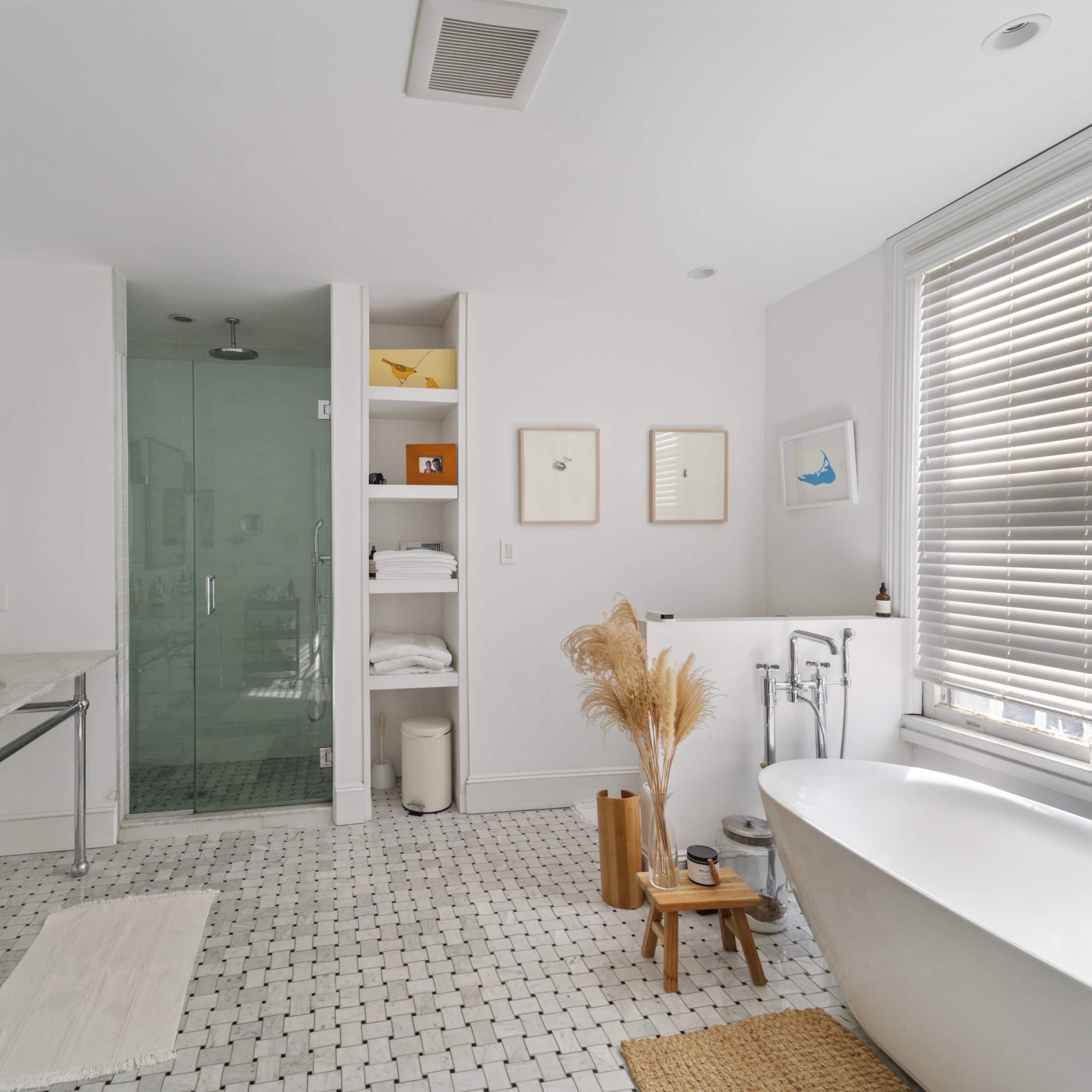 A modern bathroom features a freestanding bathtub, a walk-in shower with glass doors, a double vanity with a marble countertop, and decorative items such as a small plant and towels on shelves.