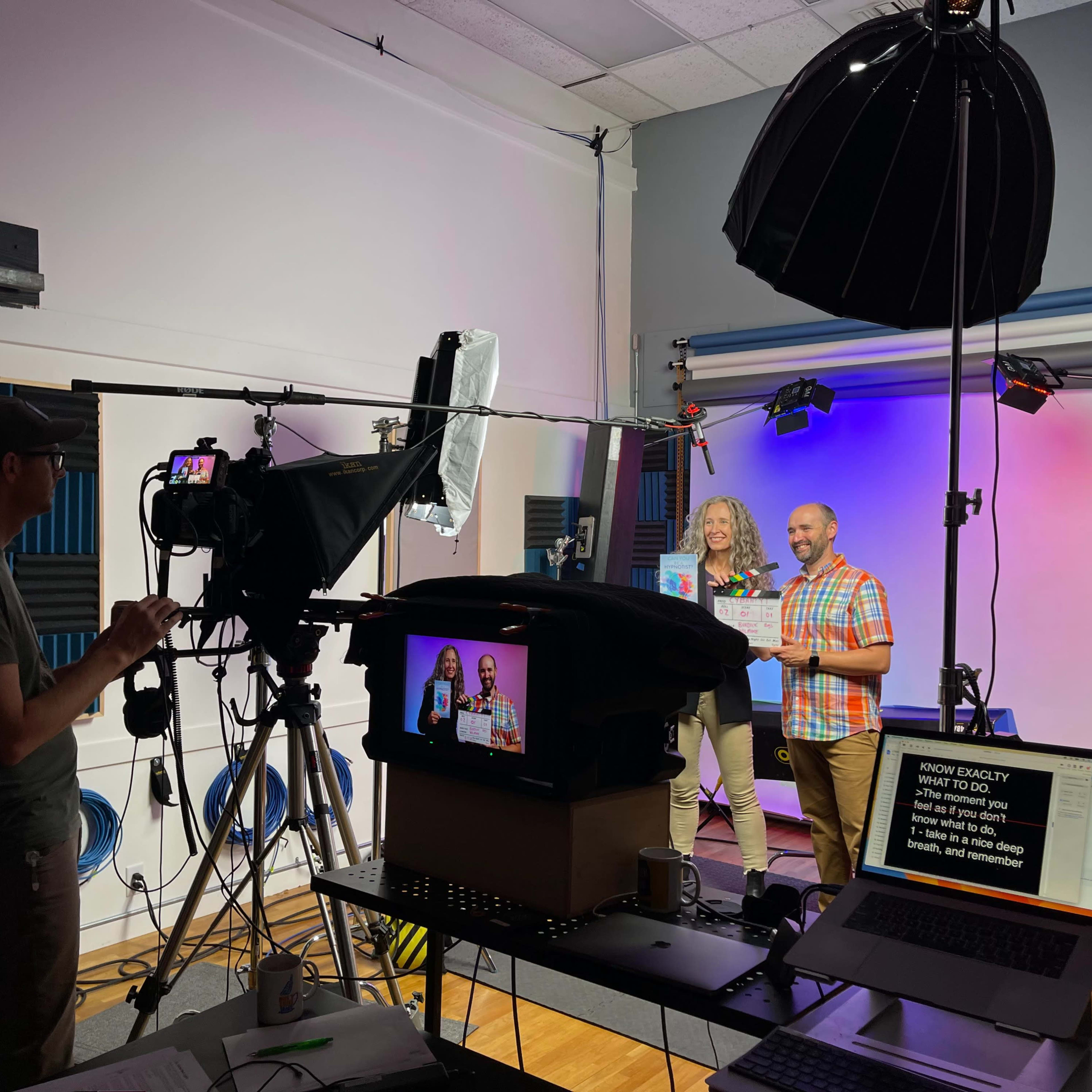 A video production crew sets up equipment while two people hold awards in front of a colorful backdrop.