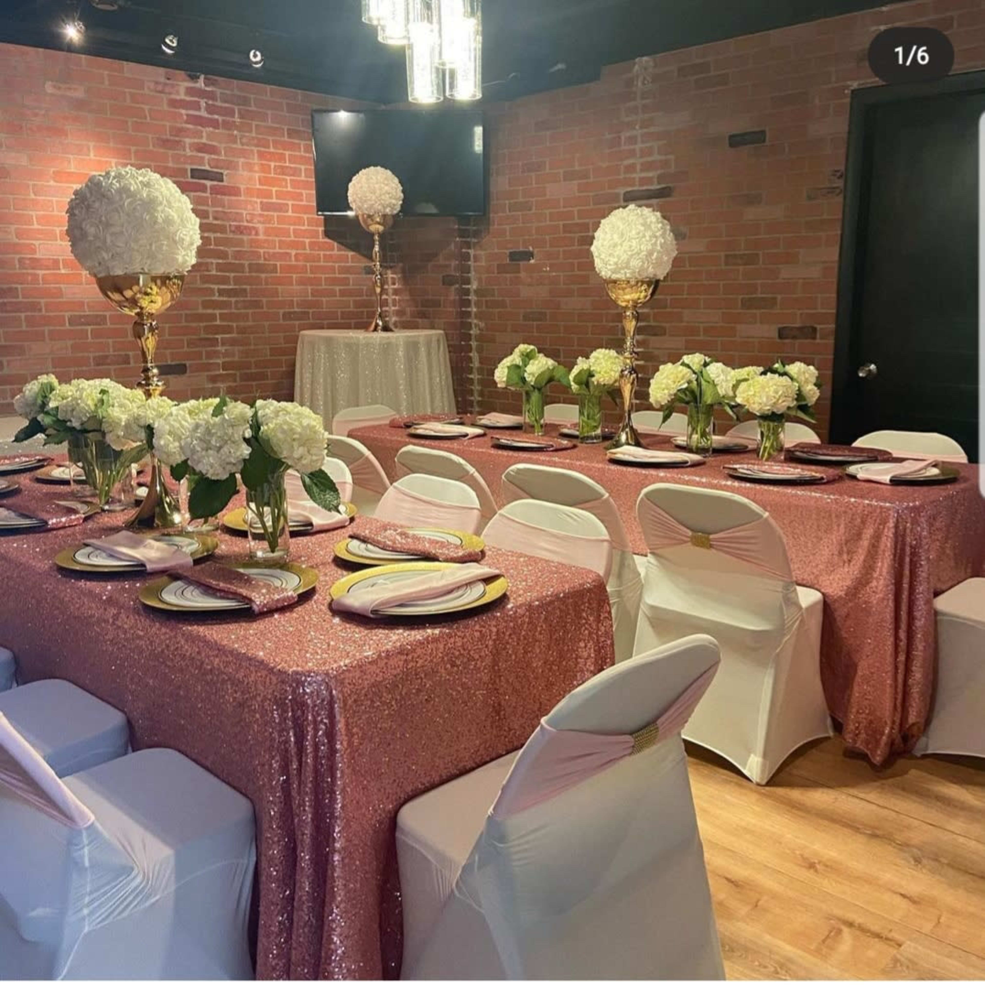 A banquet setting with multiple tables draped in pink sequin tablecloths, adorned with floral centerpieces and elegant white chair covers, in a room featuring exposed brick walls and modern lighting.