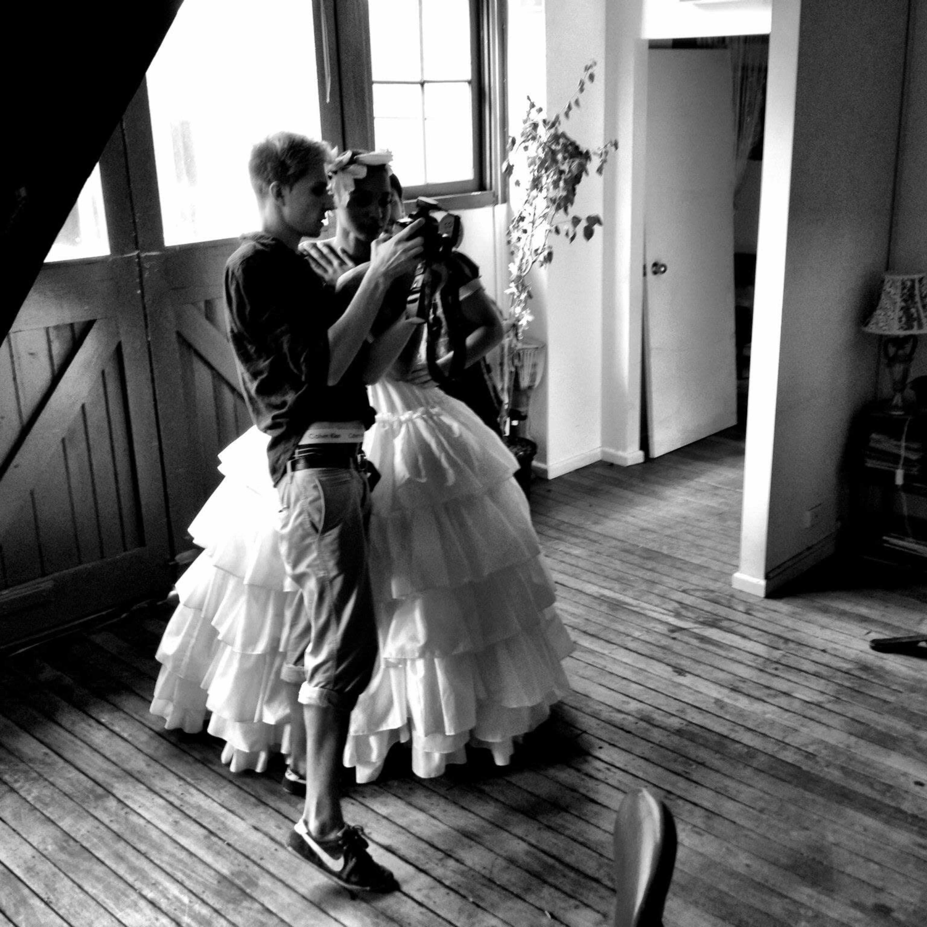 A young man is inspecting a camera while standing next to a woman in a long, ruffled white skirt inside a room with wooden floors and large windows.