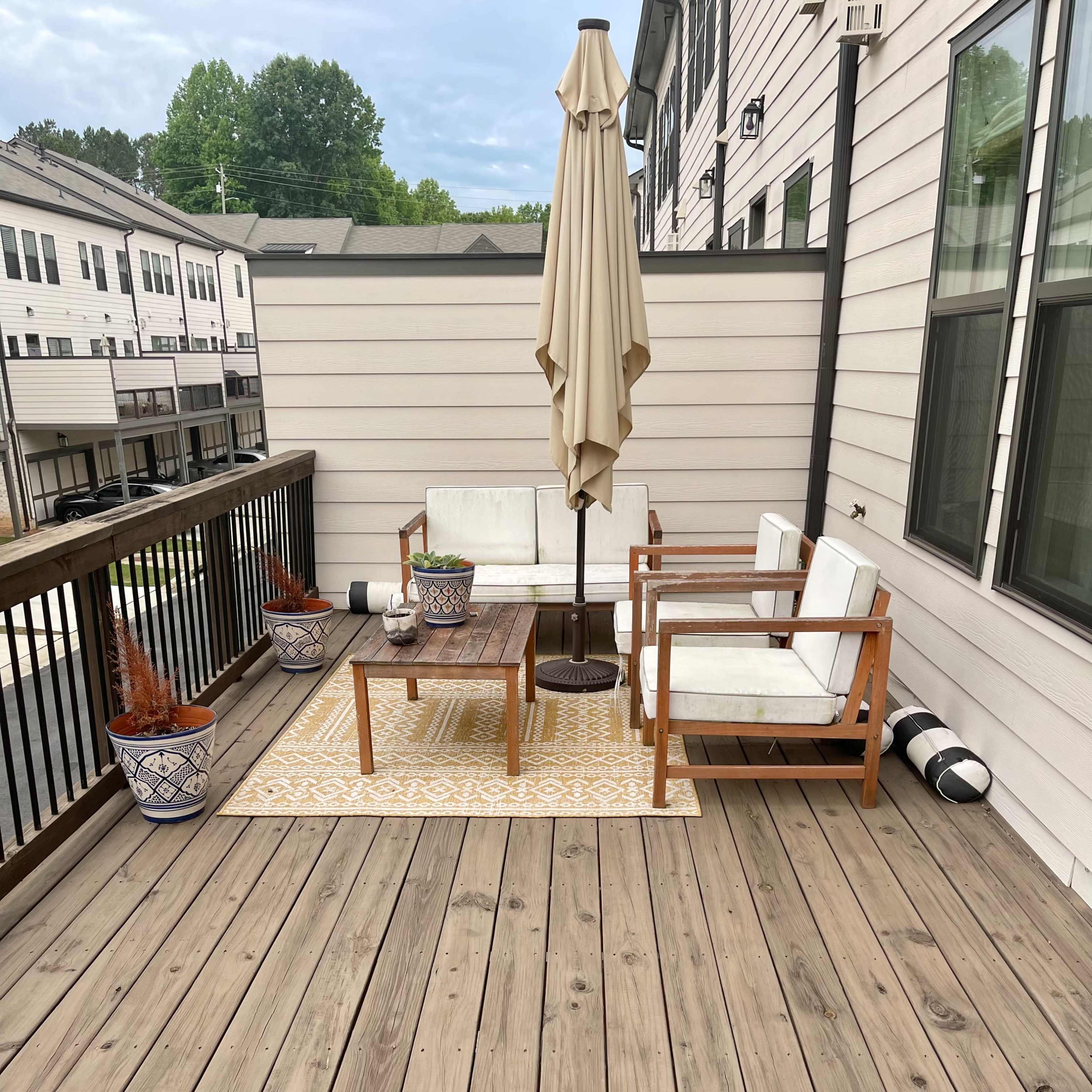 A wooden deck features two chairs, a table, an umbrella, and decorative pots, with a patterned rug in the center.