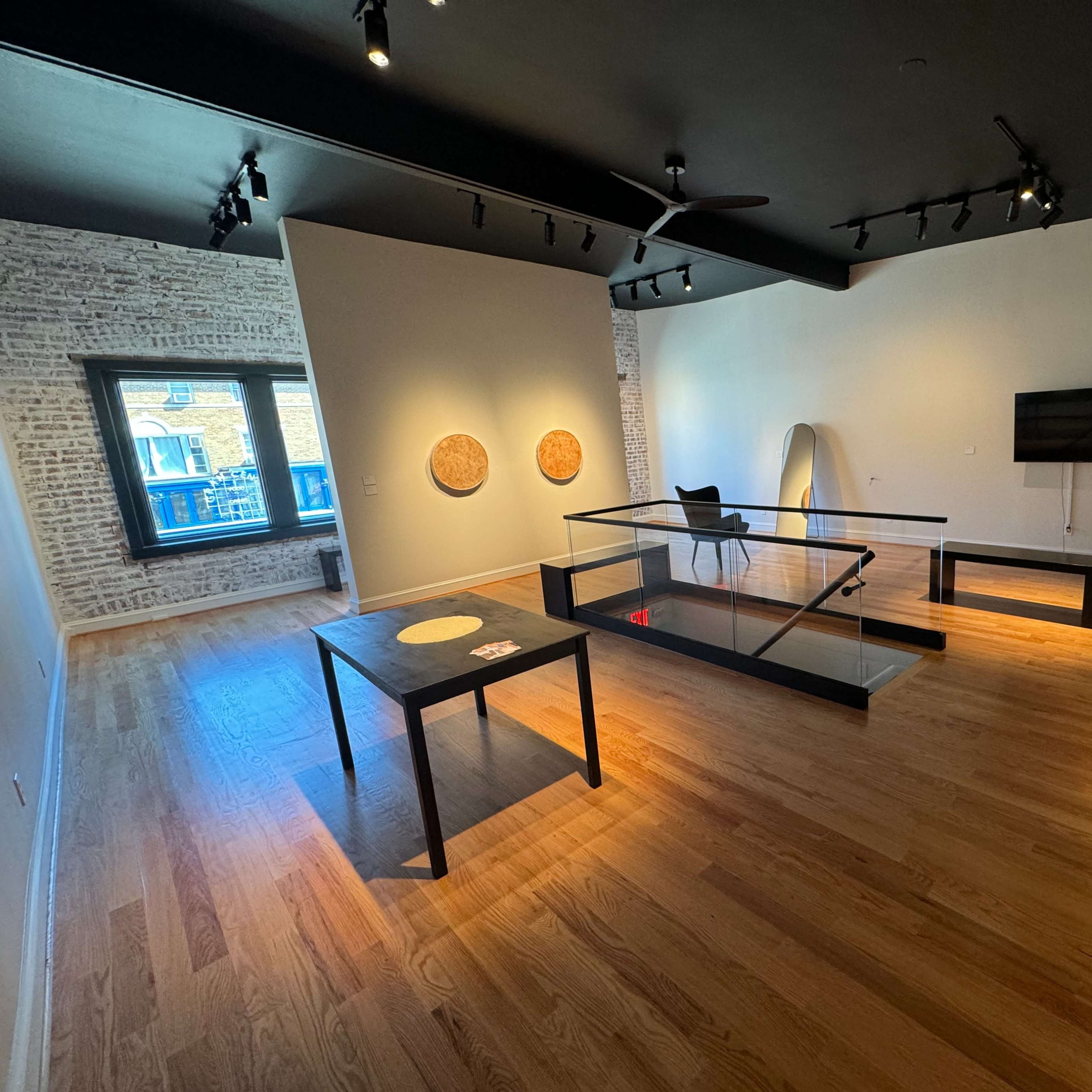 A spacious, modern room with wooden flooring, featuring a large glass table, a chair, and two circular artworks on the walls.