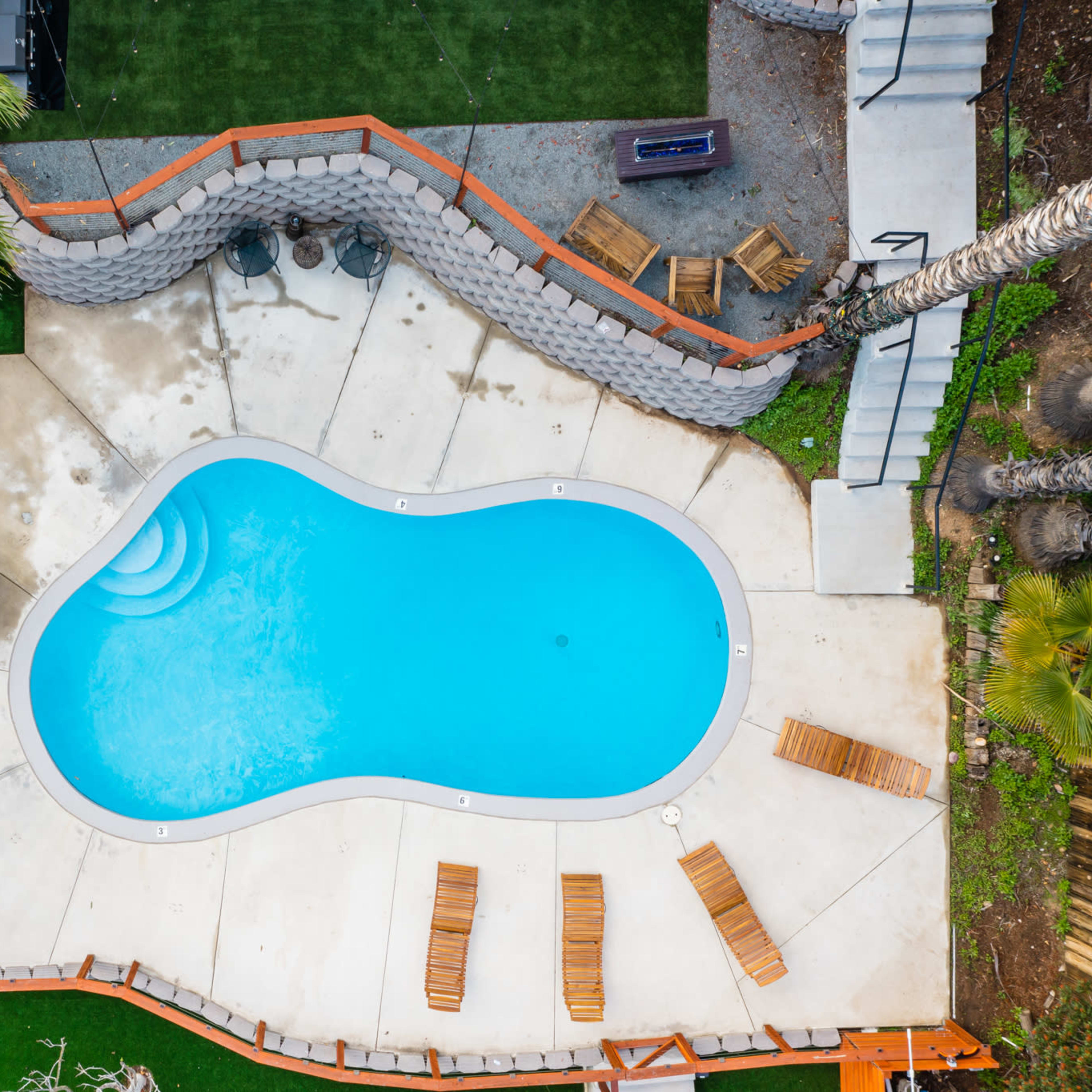 An aerial view shows an irregularly shaped swimming pool surrounded by concrete, lounge chairs, a fire pit, and decorative landscaping.
