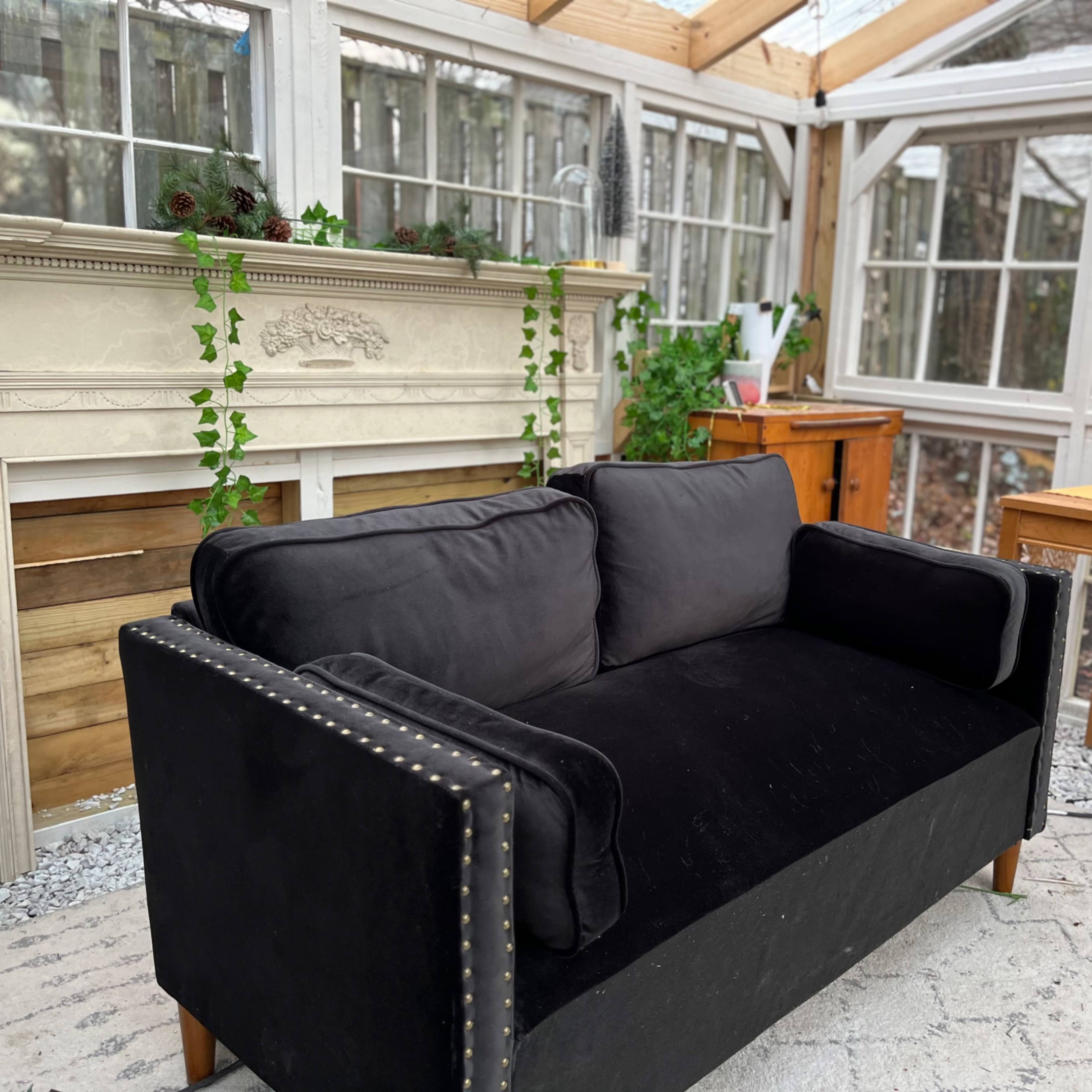 A black couch with decorative nails sits in a bright sunroom featuring large windows and a mantelpiece decorated with greenery.