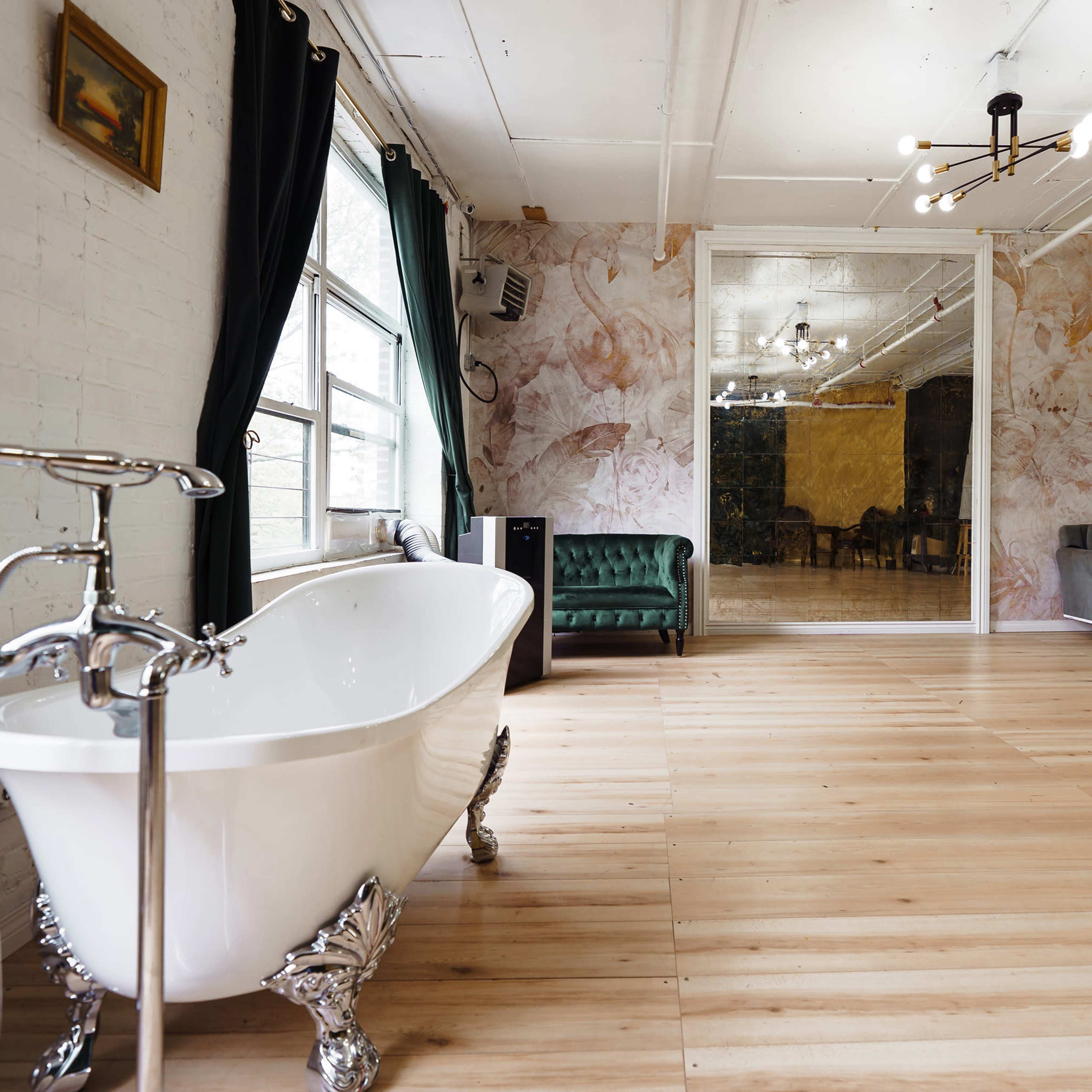 A spacious room with a vintage clawfoot bathtub, a green velvet chair, a gray sofa, and a mirrored wall.