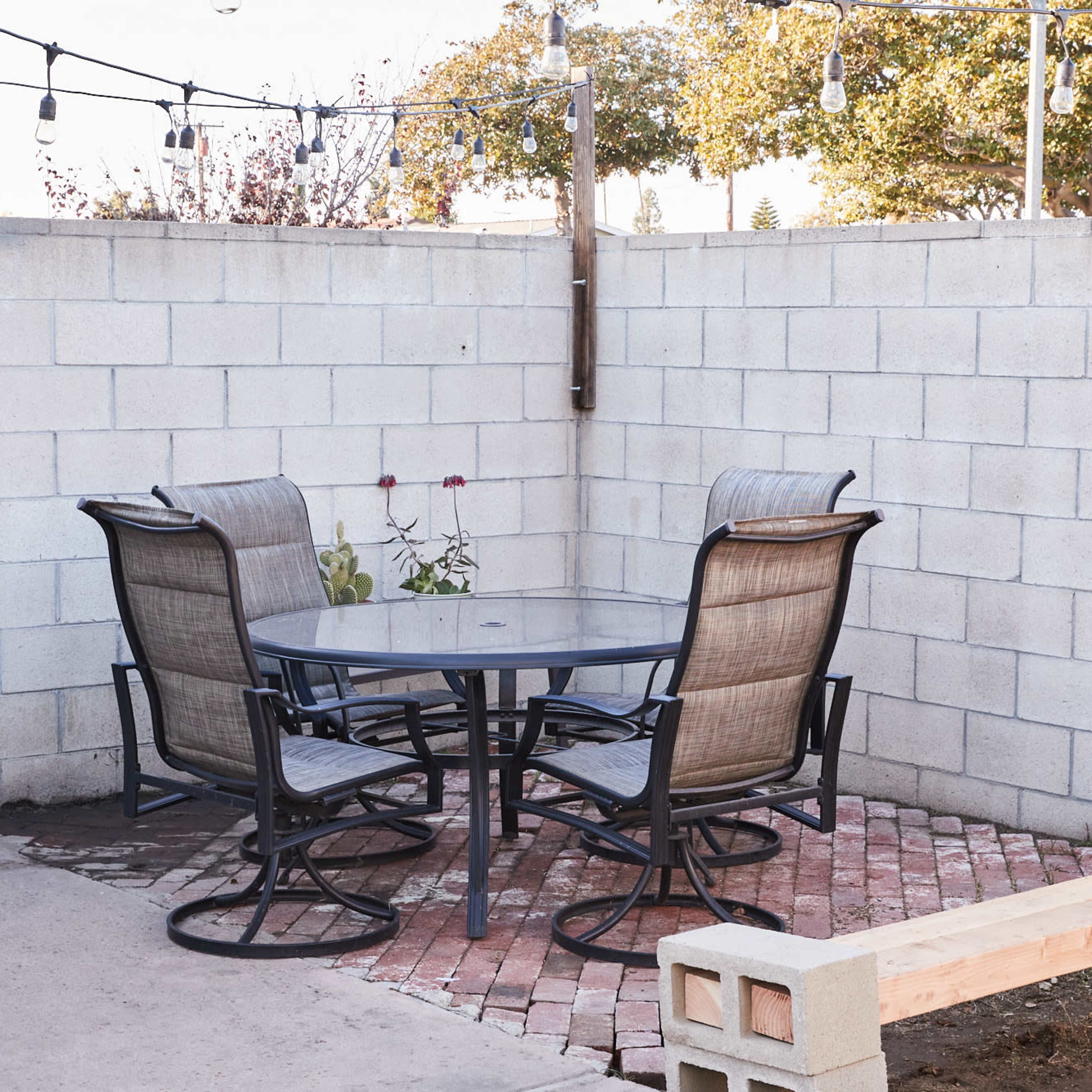 A small outdoor seating area features a round glass table surrounded by four metal chairs on a brick patio enclosed by a cinder block wall.