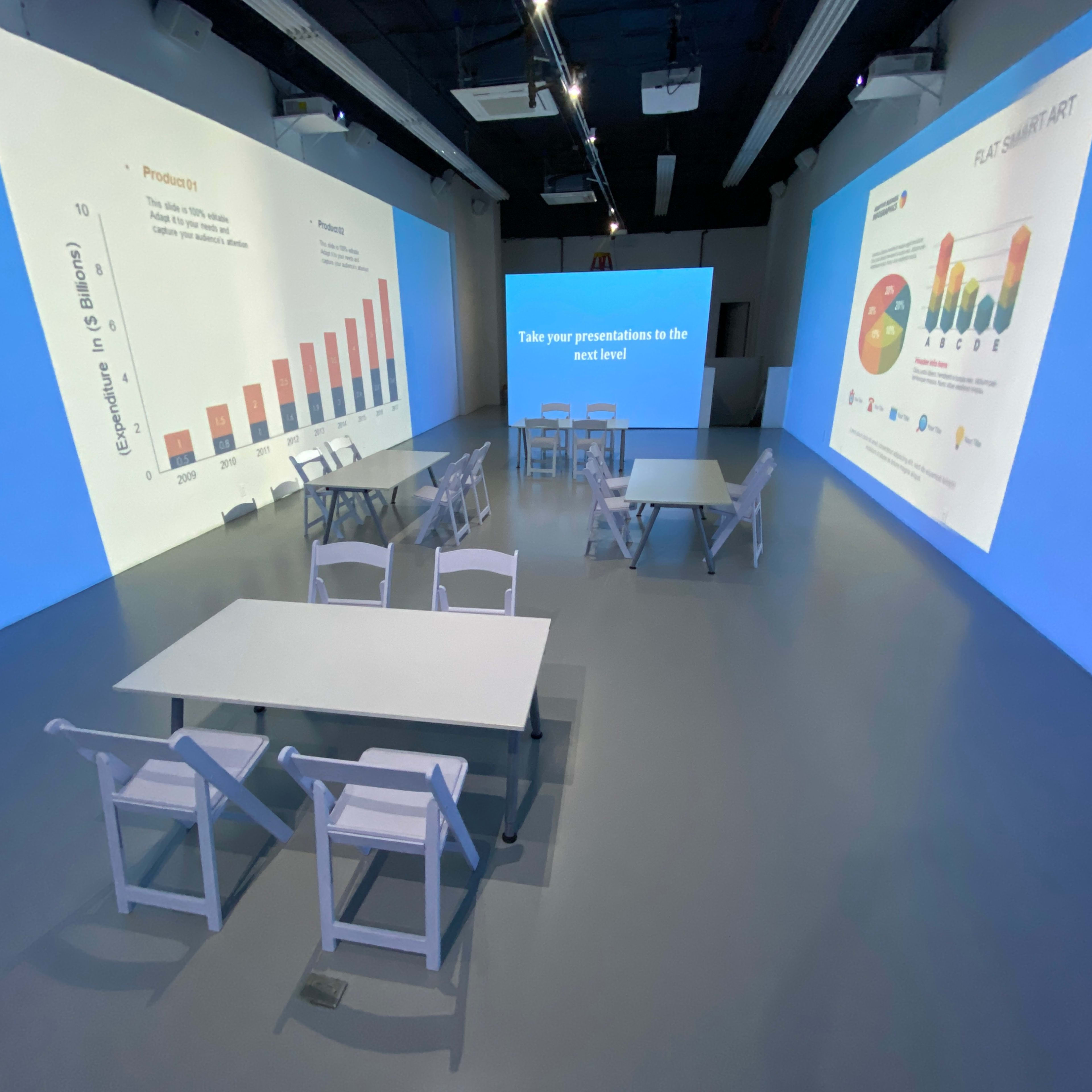 A modern presentation room with white tables and chairs, featuring large screens displaying graphs and charts on the walls.