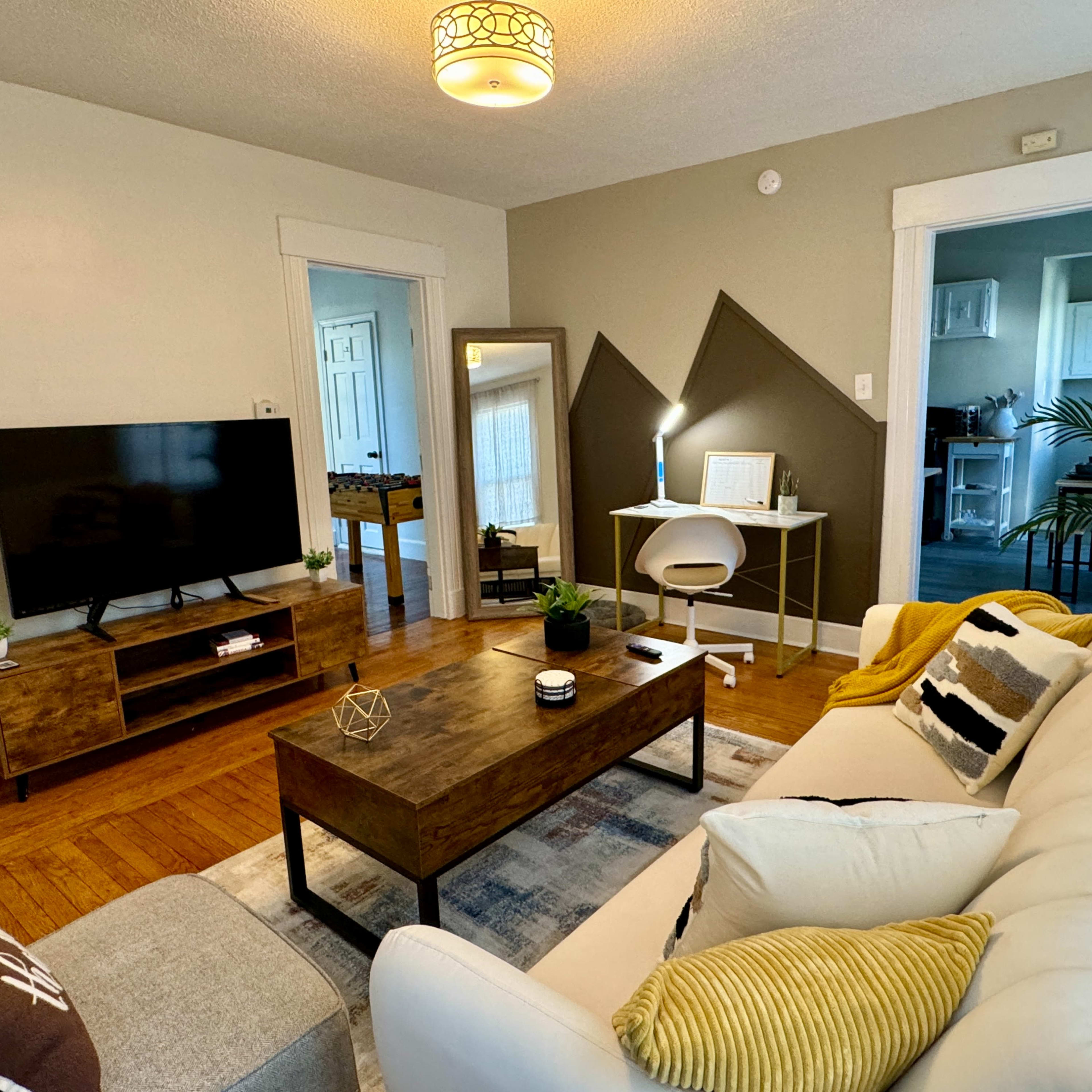 The living room features a white sofa, a wooden coffee table, a television on a low cabinet, and a workspace with a chair and lamp against a backdrop of light-colored walls and angles.