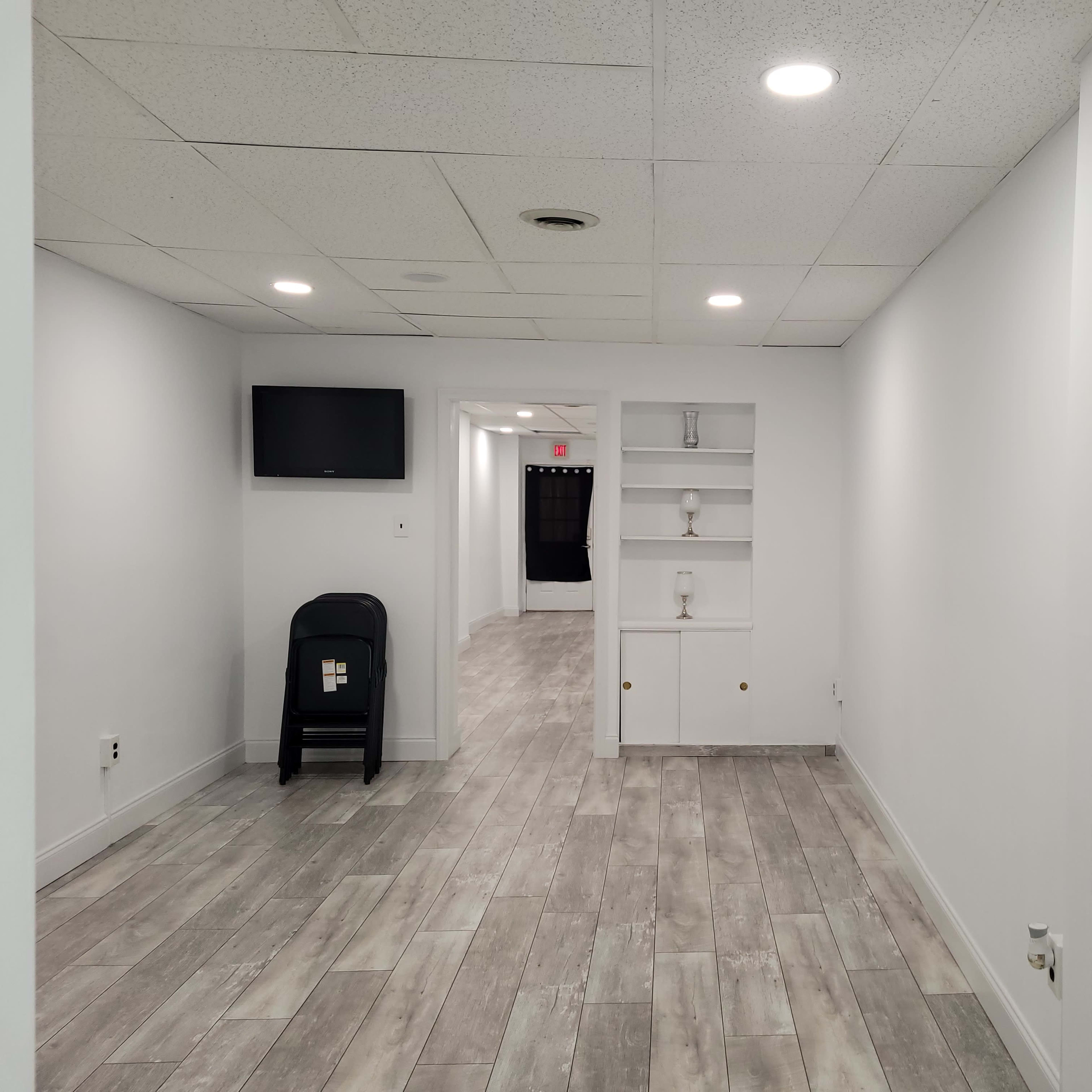 A well-lit, empty room with light-colored walls and flooring, featuring a TV mounted on the wall and a small shelf unit.