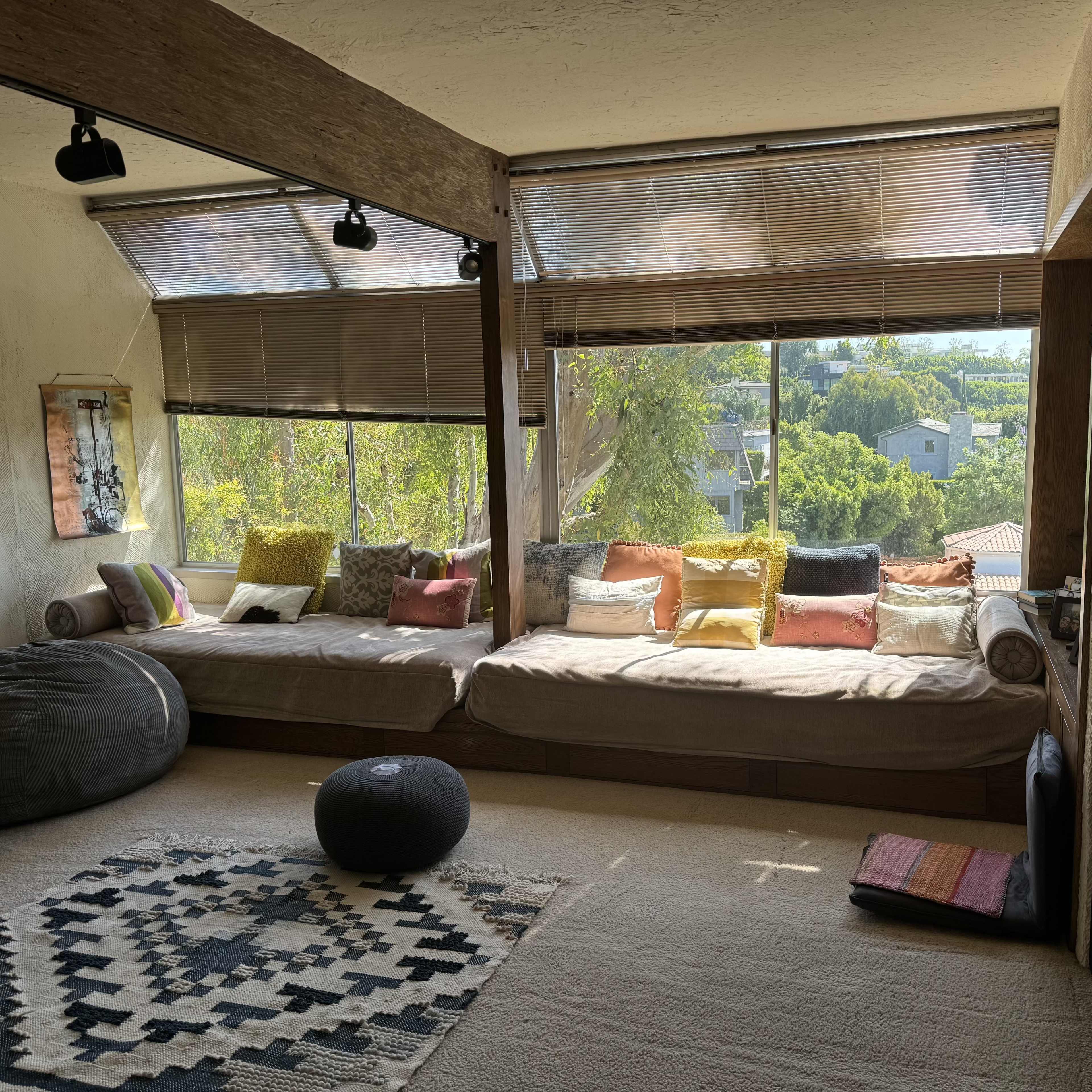A cozy living area with large windows, featuring a low seating area adorned with colorful pillows and a patterned rug on a carpeted floor.