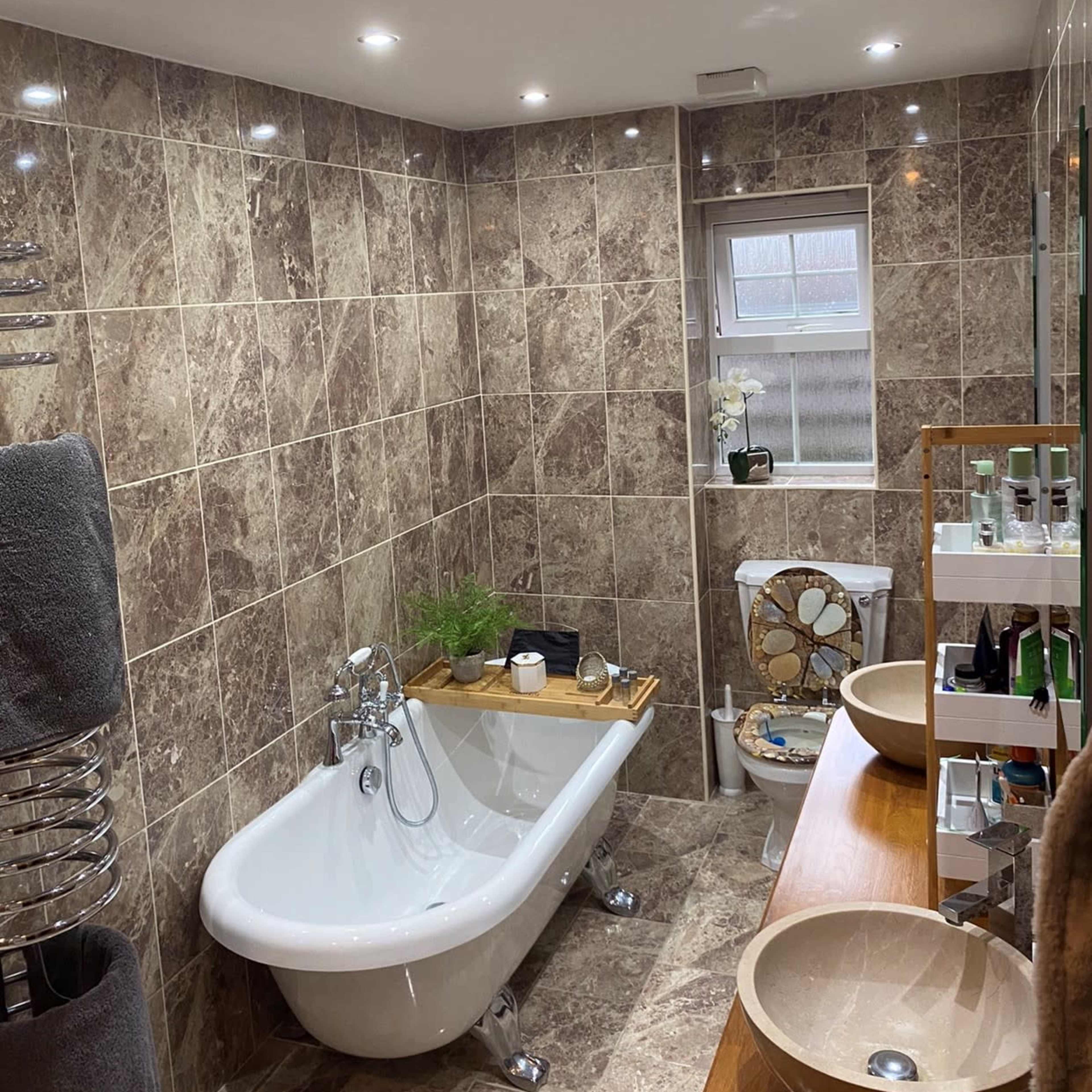 A bathroom with marble tiled walls, a freestanding bathtub, two sinks on a wooden countertop, and a toilet next to a window.