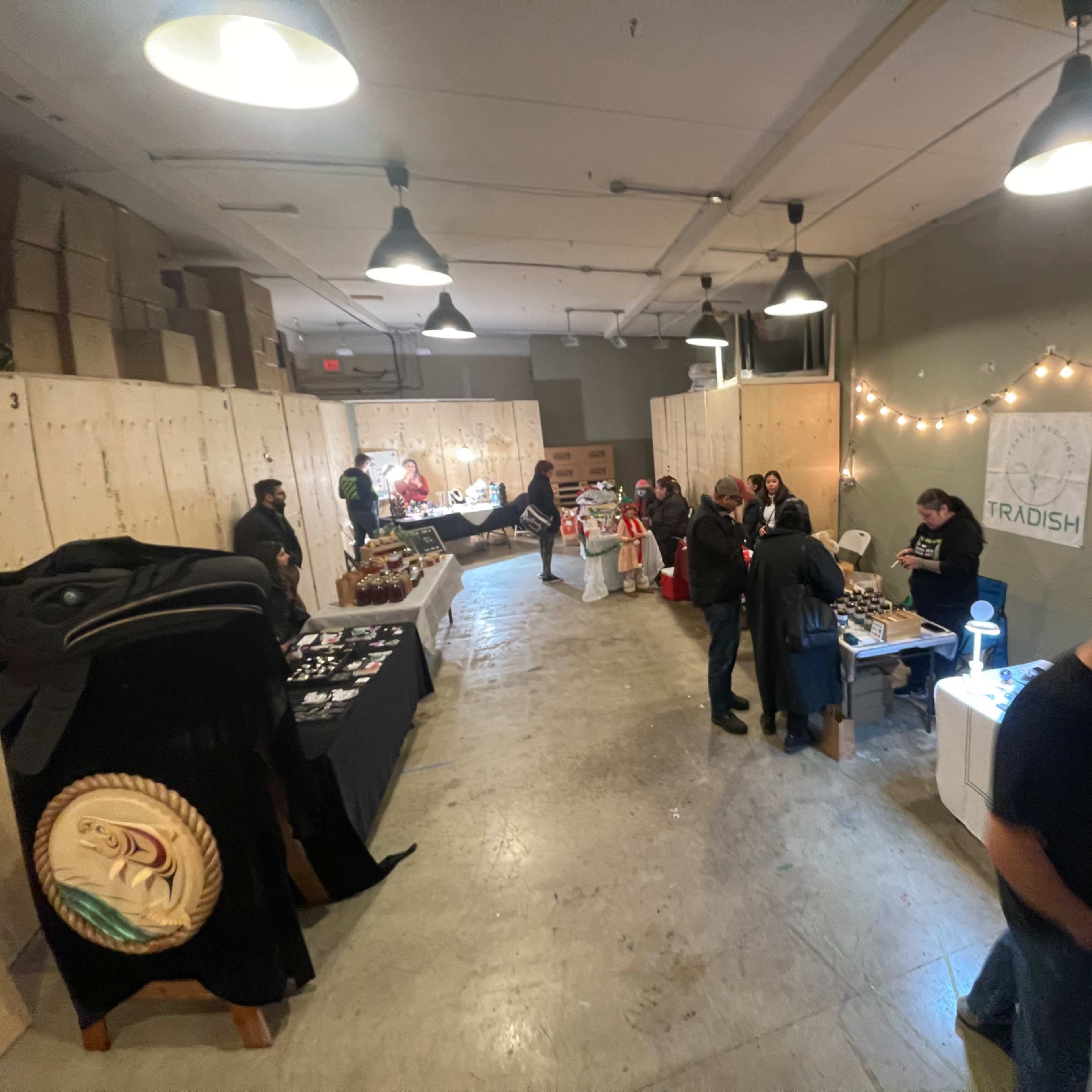 A crowded indoor marketplace features various tables displaying products, with shoppers interacting and browsing among the stalls.