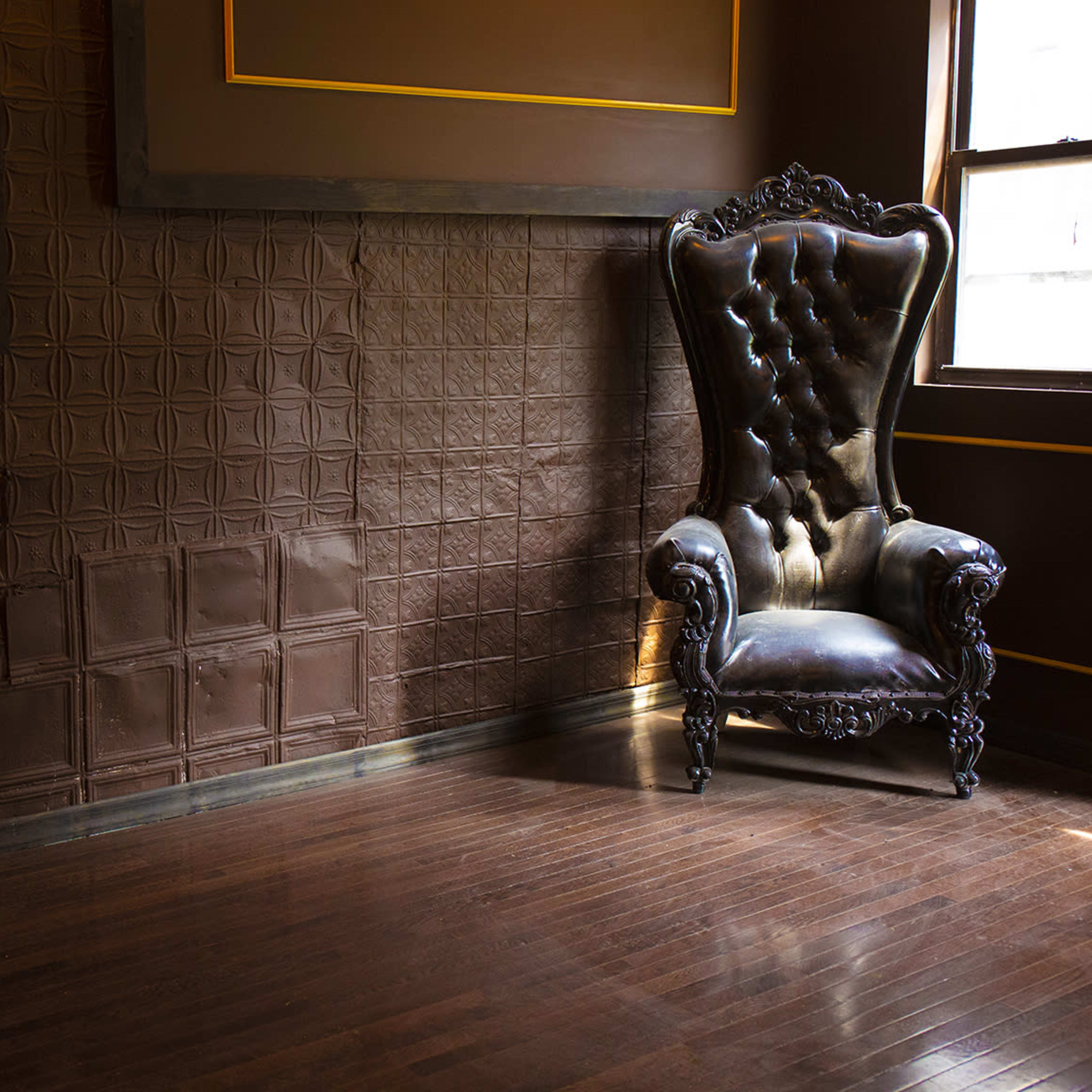 A large, ornate black chair is positioned against a wall with patterned panels and gold trim in a dimly lit room.