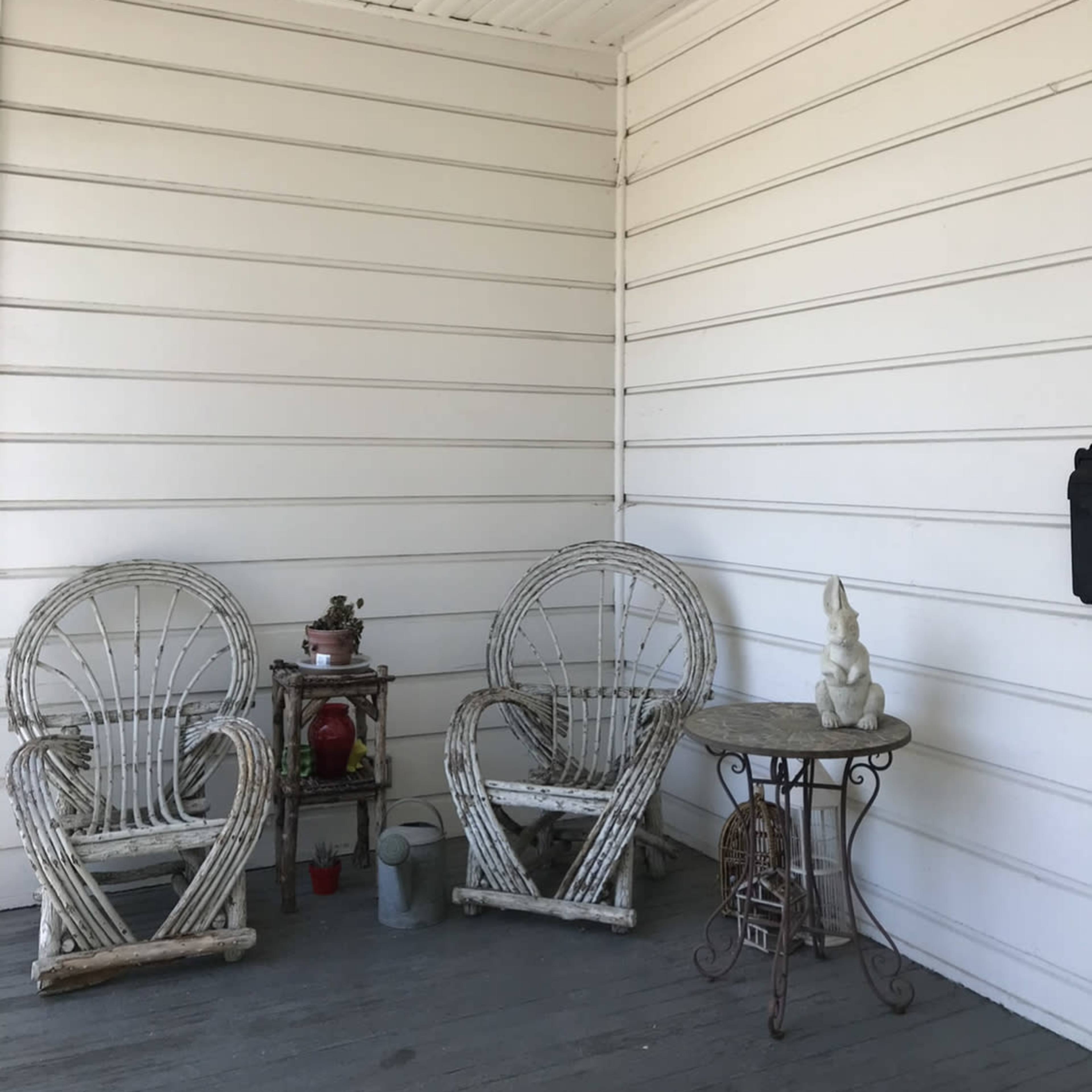 A porch with two wicker chairs facing a small table and a decorative sculpture against a white-paneled wall.