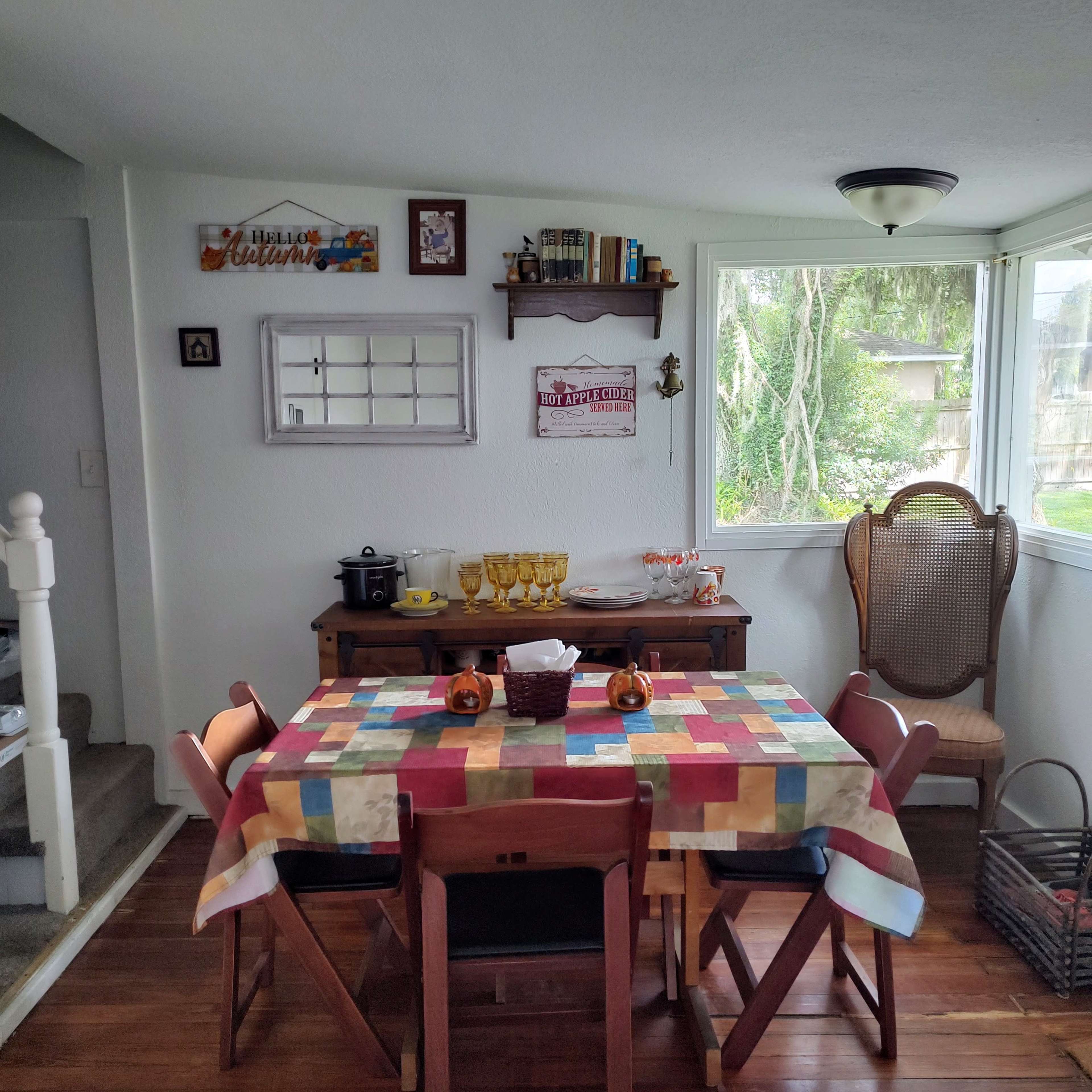 A dining area with a colorful tablecloth-covered table surrounded by wooden chairs, a sideboard with various dishes, and large windows letting in natural light.