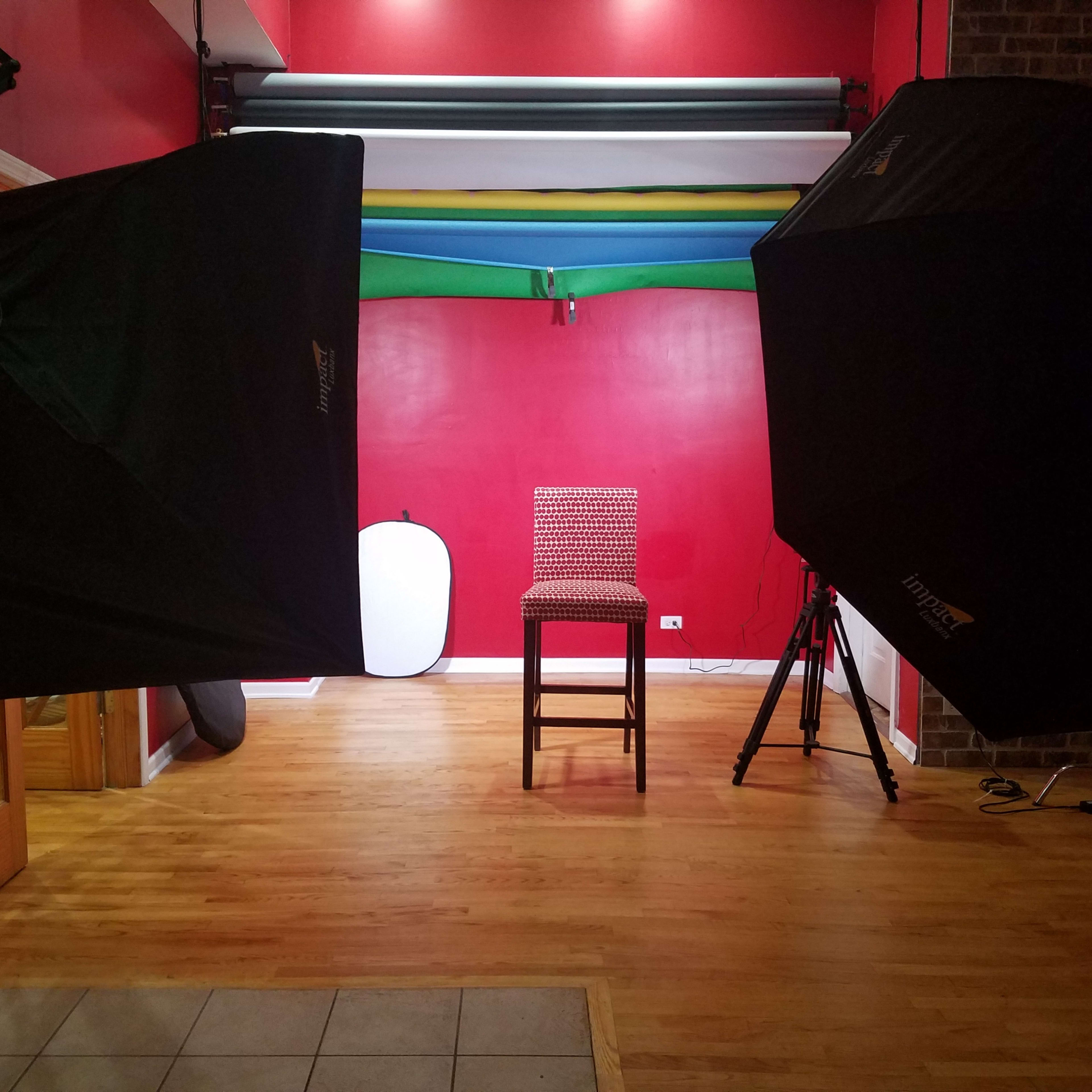 A photography studio with two large softbox lights facing a red wall, featuring a single tall chair in the center.