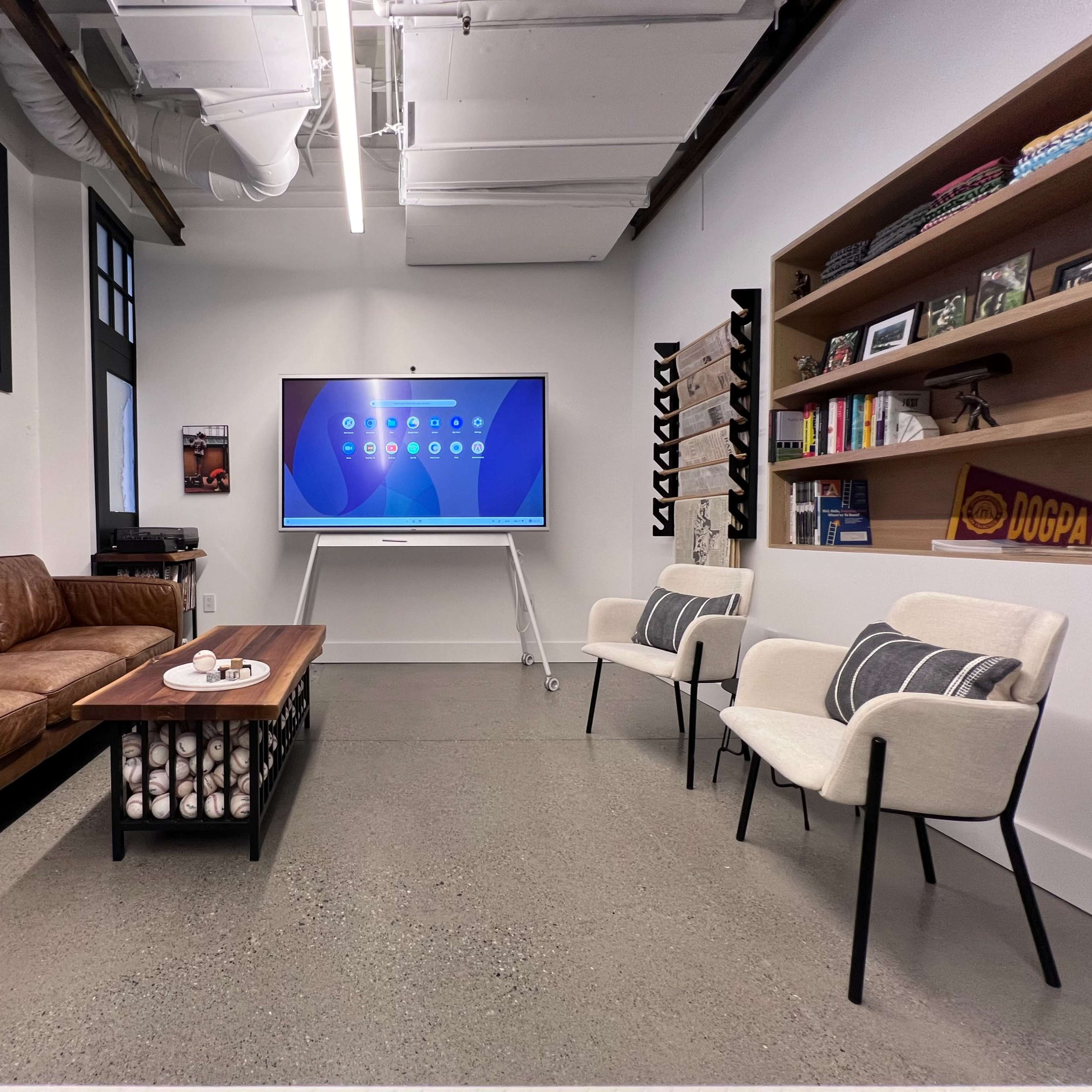 A modern office space featuring a brown leather sofa, two white chairs, a coffee table, a wall-mounted display screen, and a bookshelf against a white wall.
