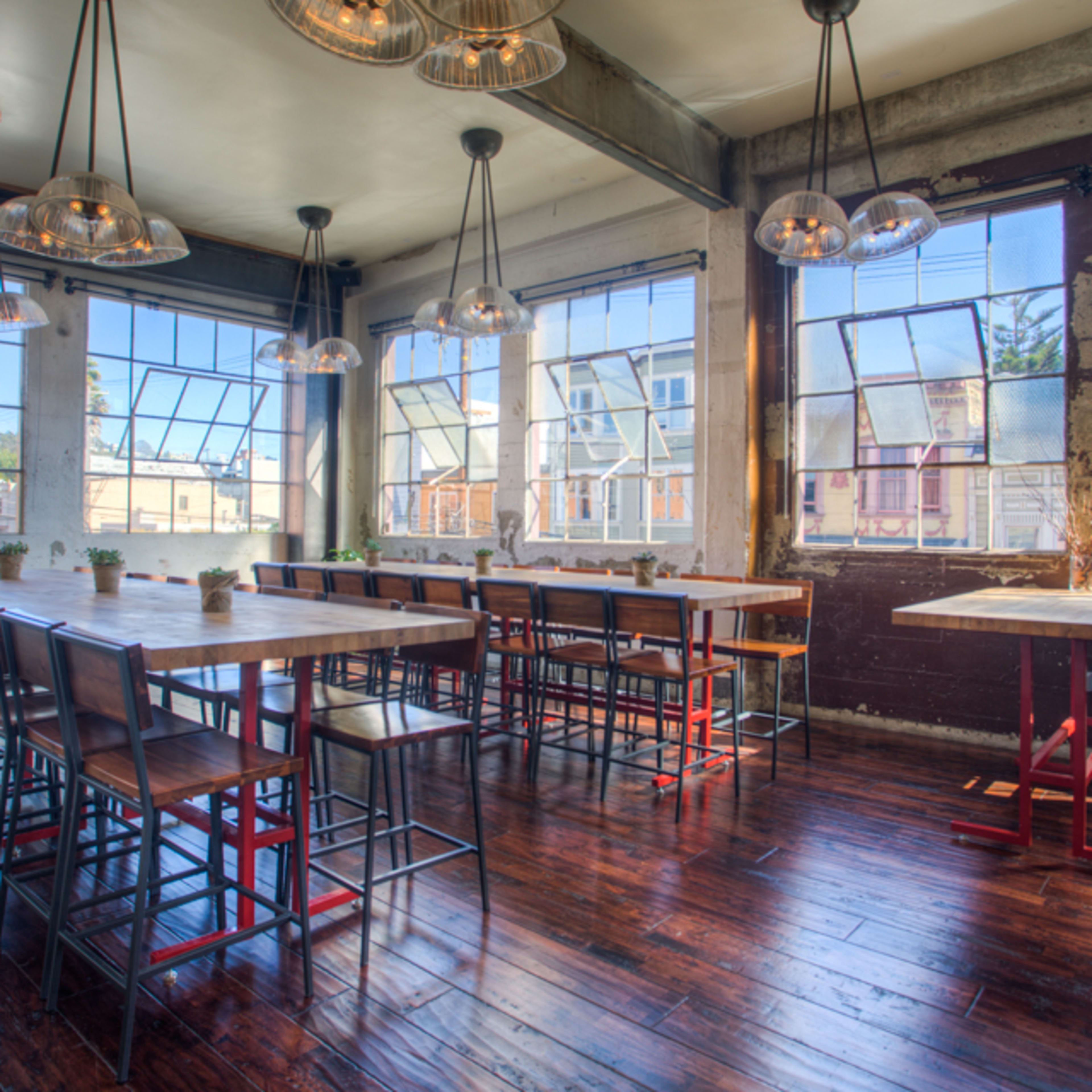 A spacious dining area with large windows, wooden floors, and long communal tables surrounded by high stools.