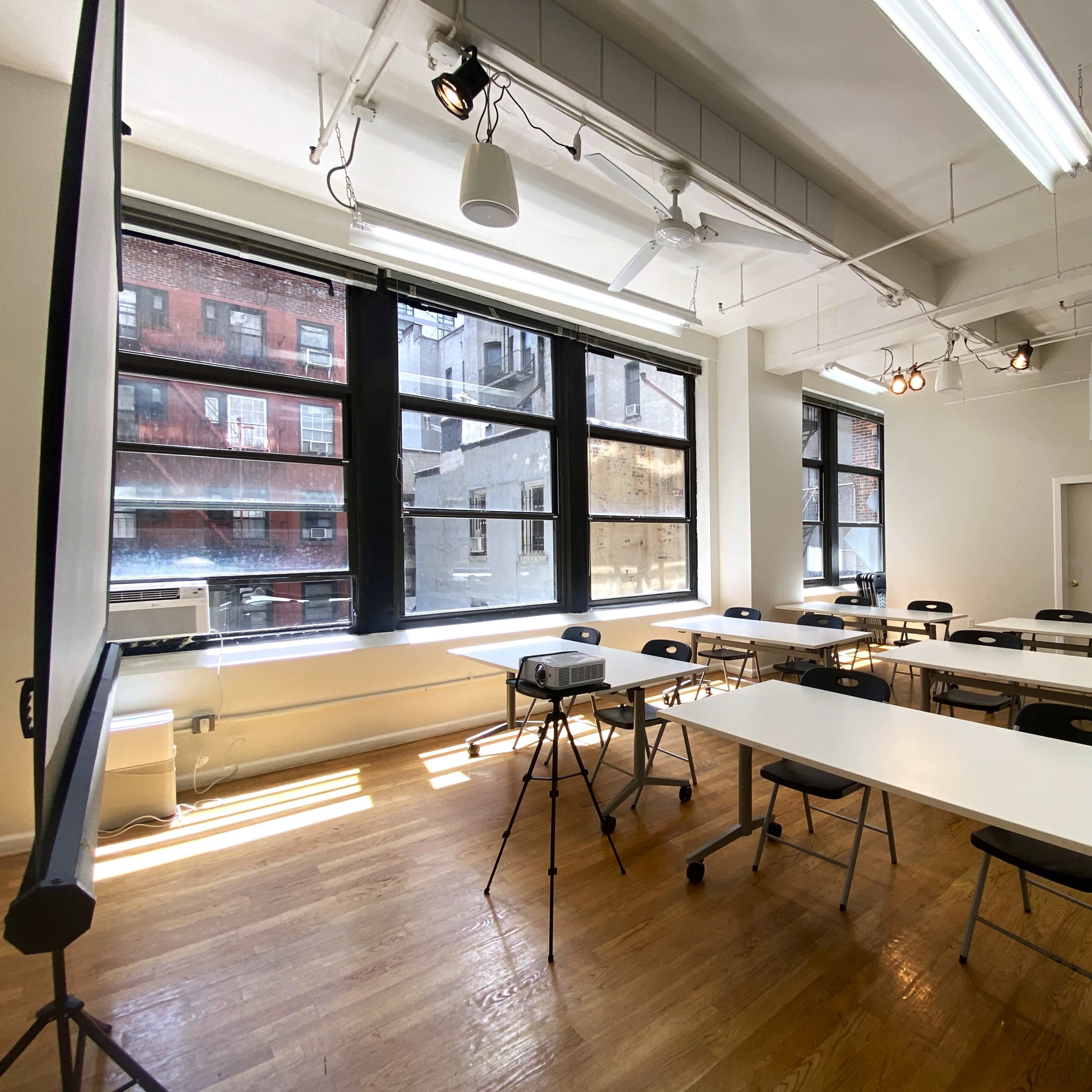 A brightly lit classroom with white tables and black chairs arranged in rows next to large windows.