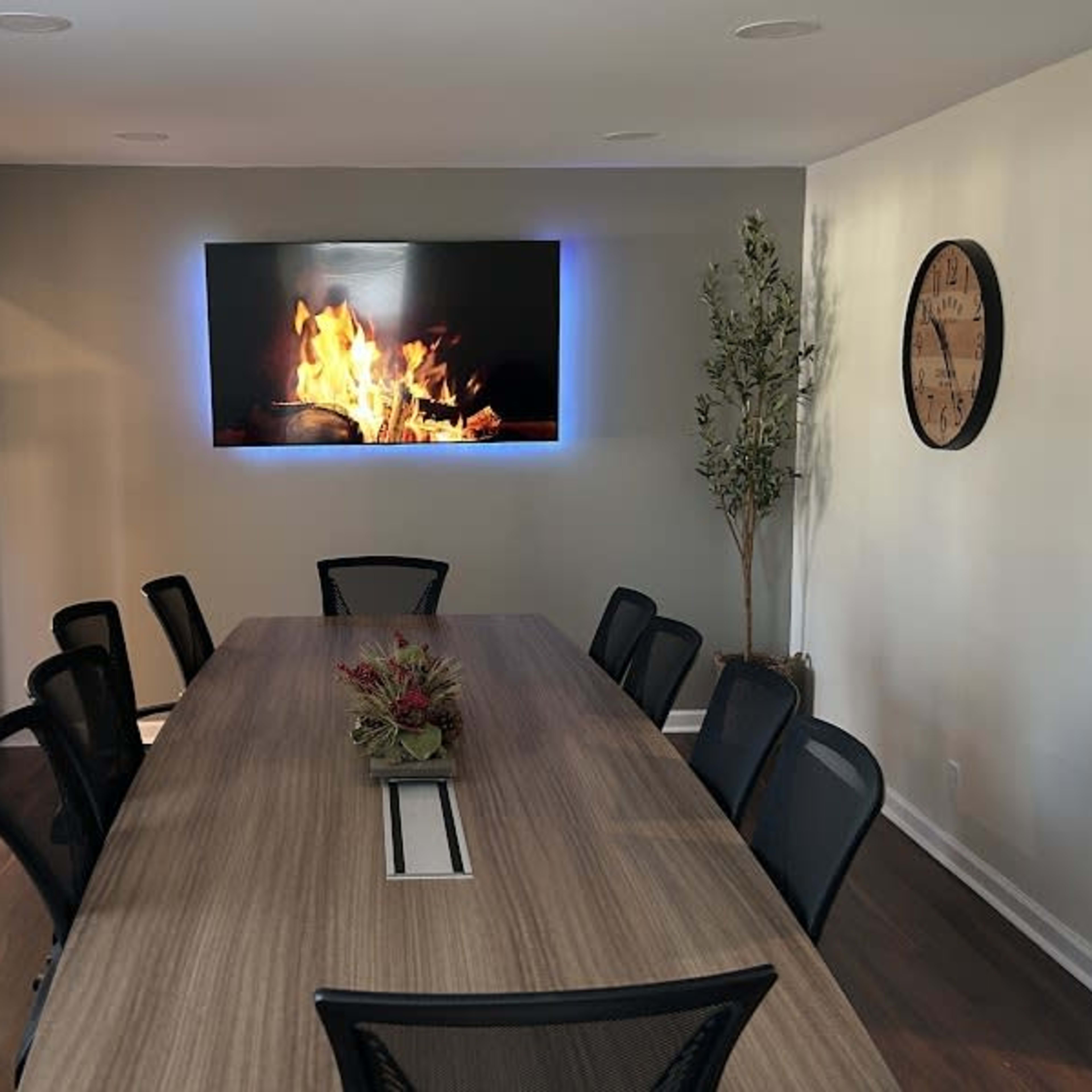 A modern conference room features a large table surrounded by black chairs, with a digital fireplace display on the wall and a decorative plant in one corner.