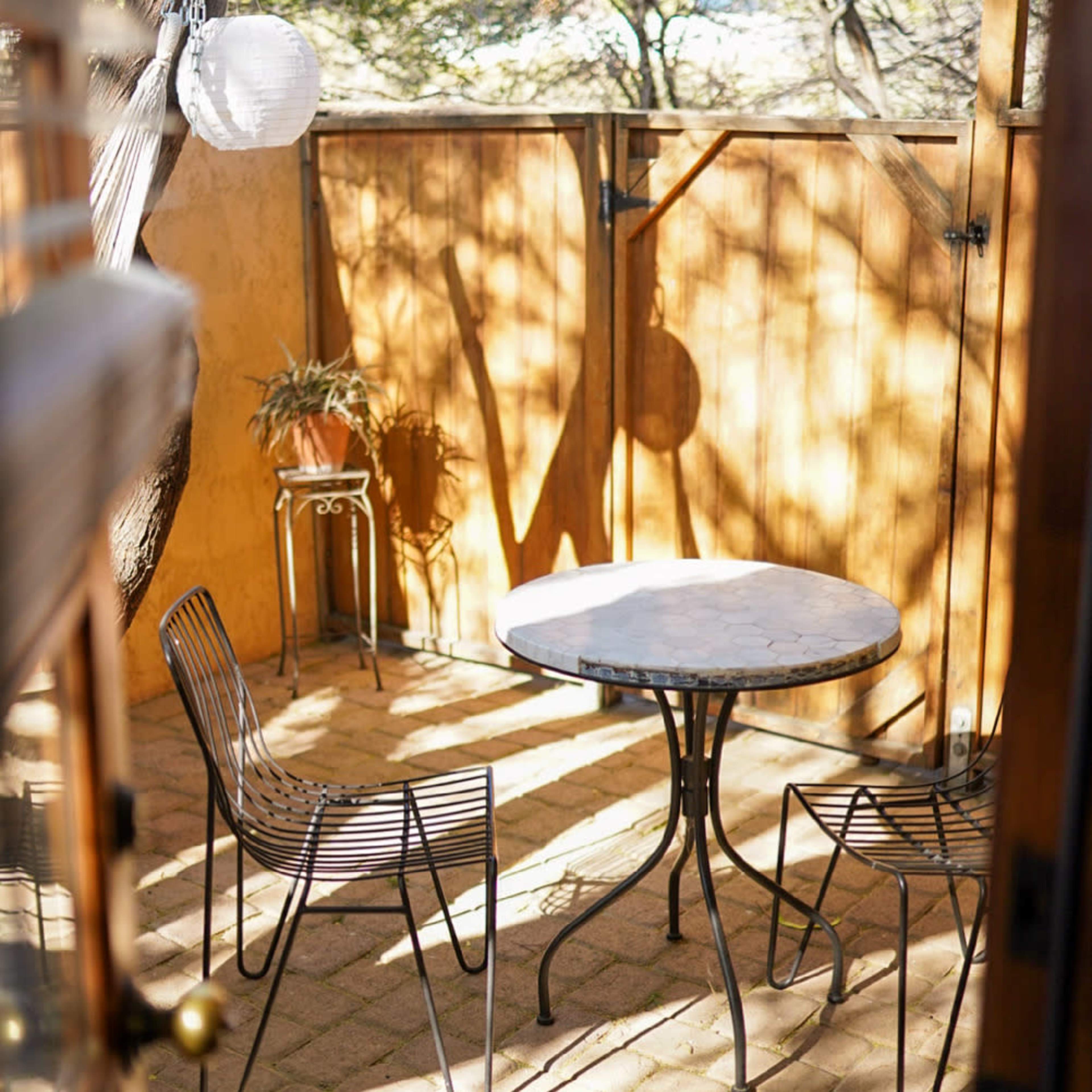 A small outdoor space features a round table with two metal chairs, surrounded by a wooden fence and dappled sunlight.
