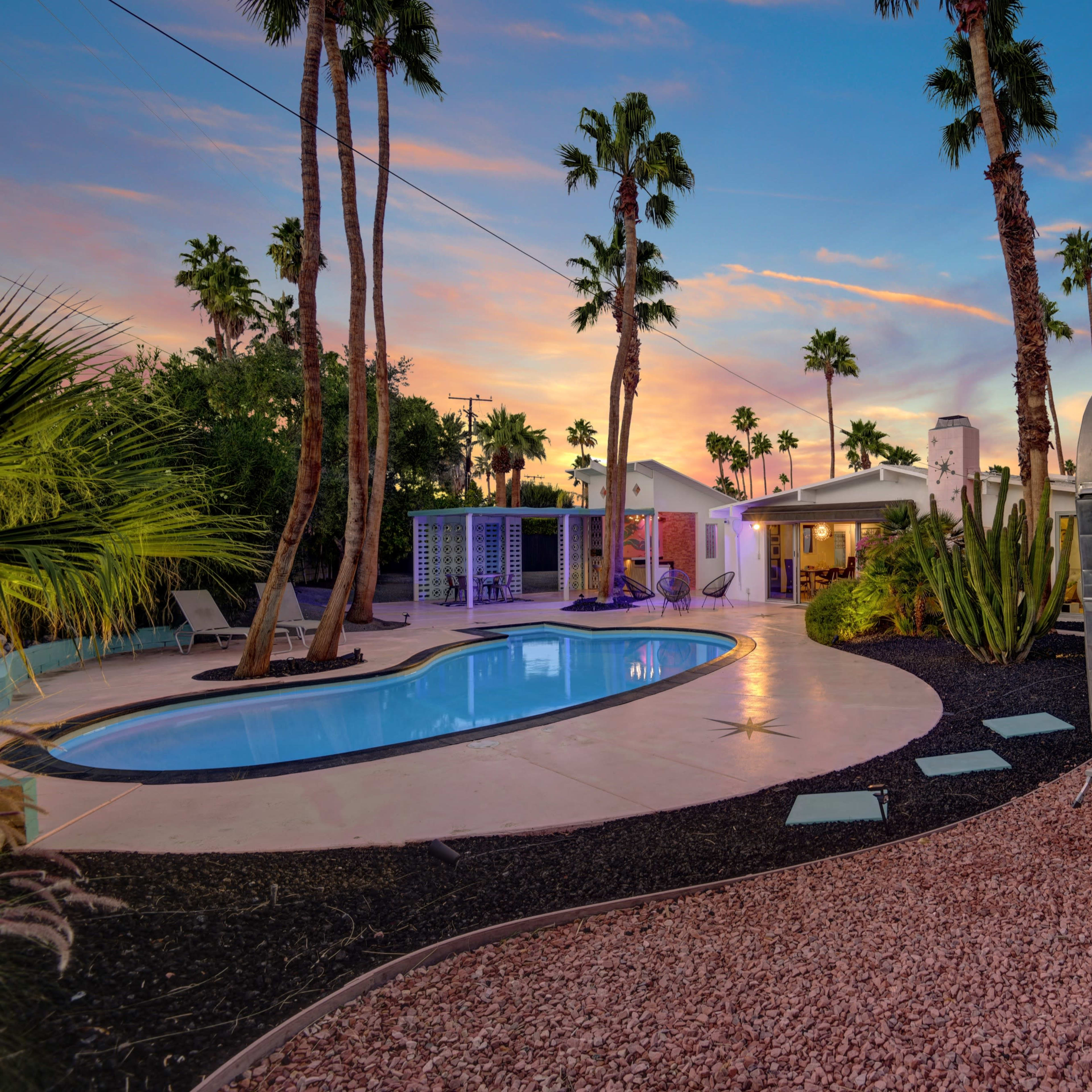 A swimming pool surrounded by a desert landscape, with palm trees and a vintage trailer in the foreground against a colorful sunset.