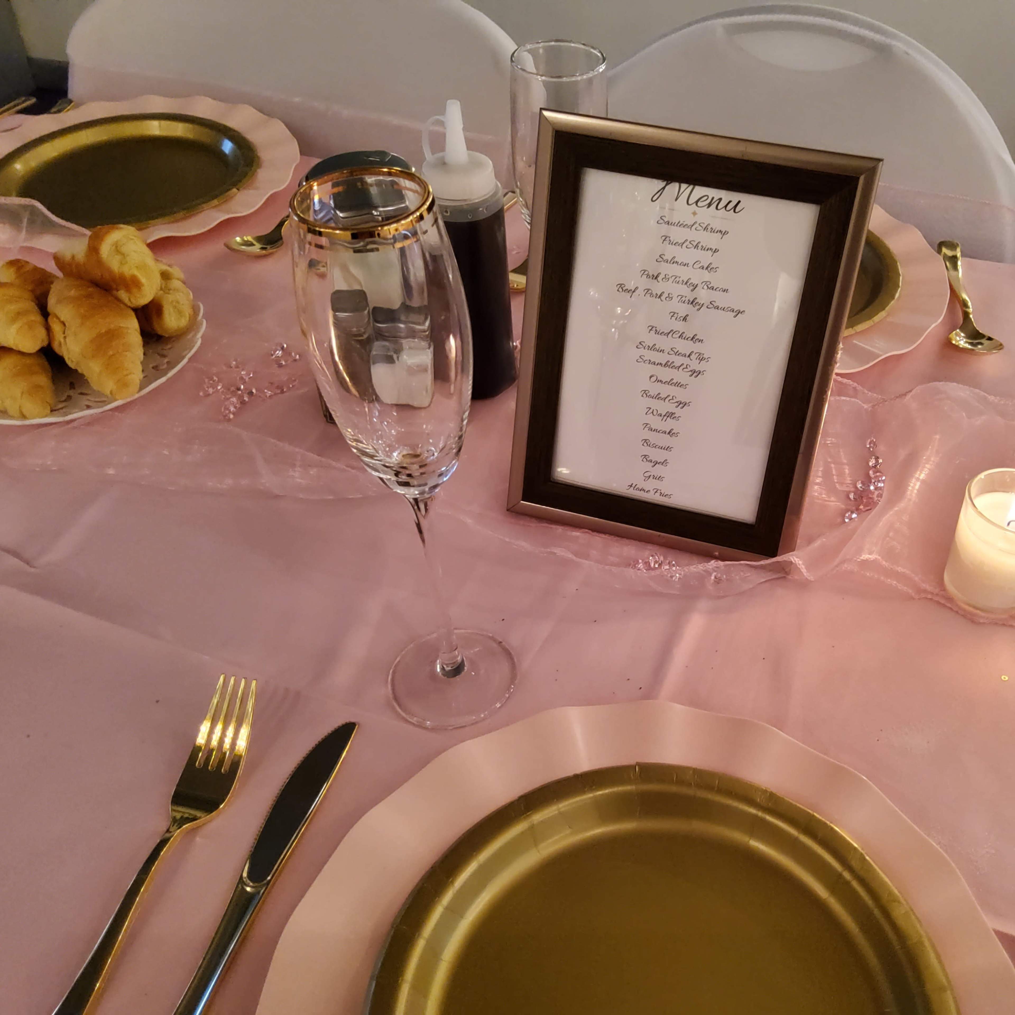 A dining table is elegantly set with gold-rimmed plates, cutlery, a framed menu, a candle, and a plate of croissants.