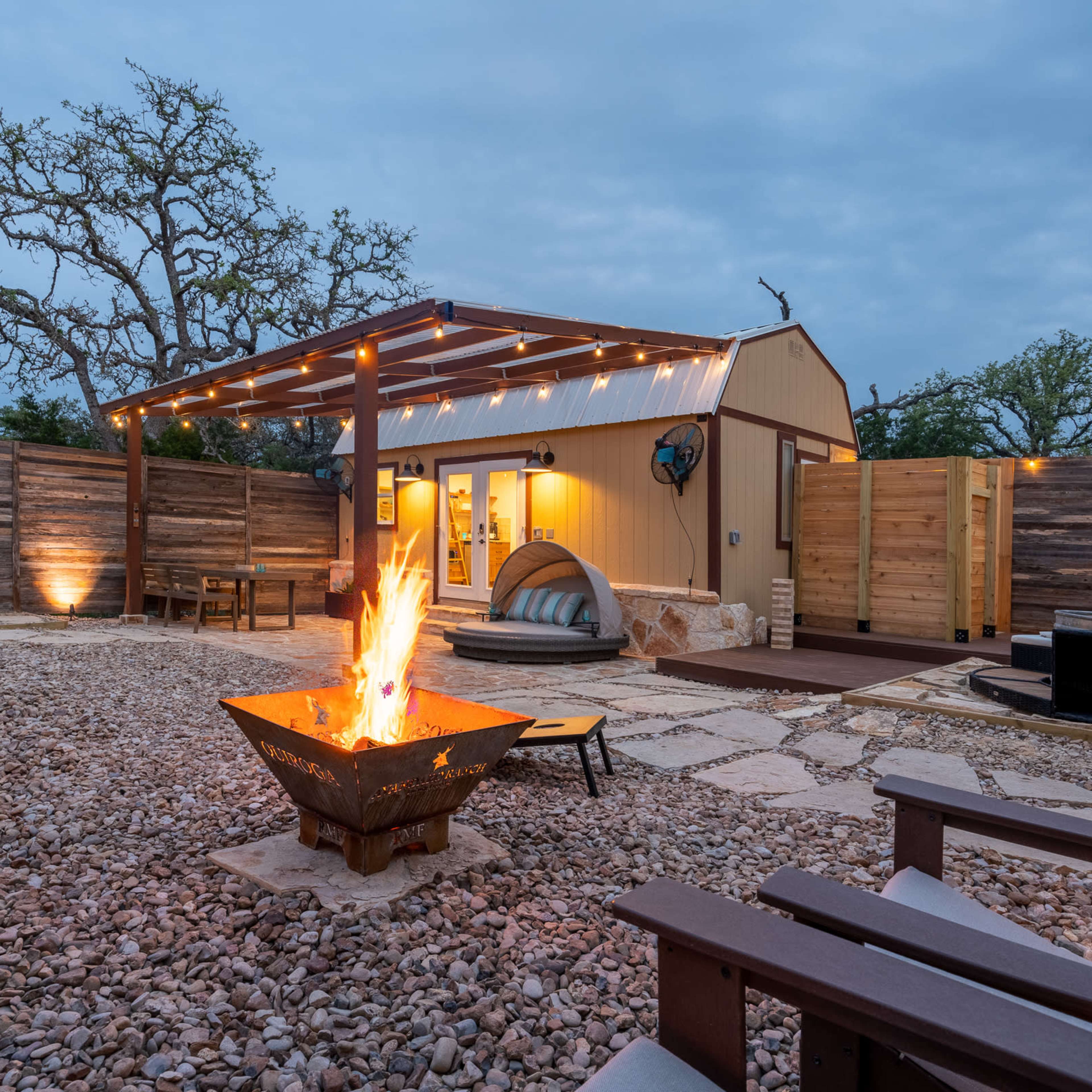 A cozy outdoor space features a fire pit surrounded by gravel, wooden seating, and a small building under a lighted pergola.