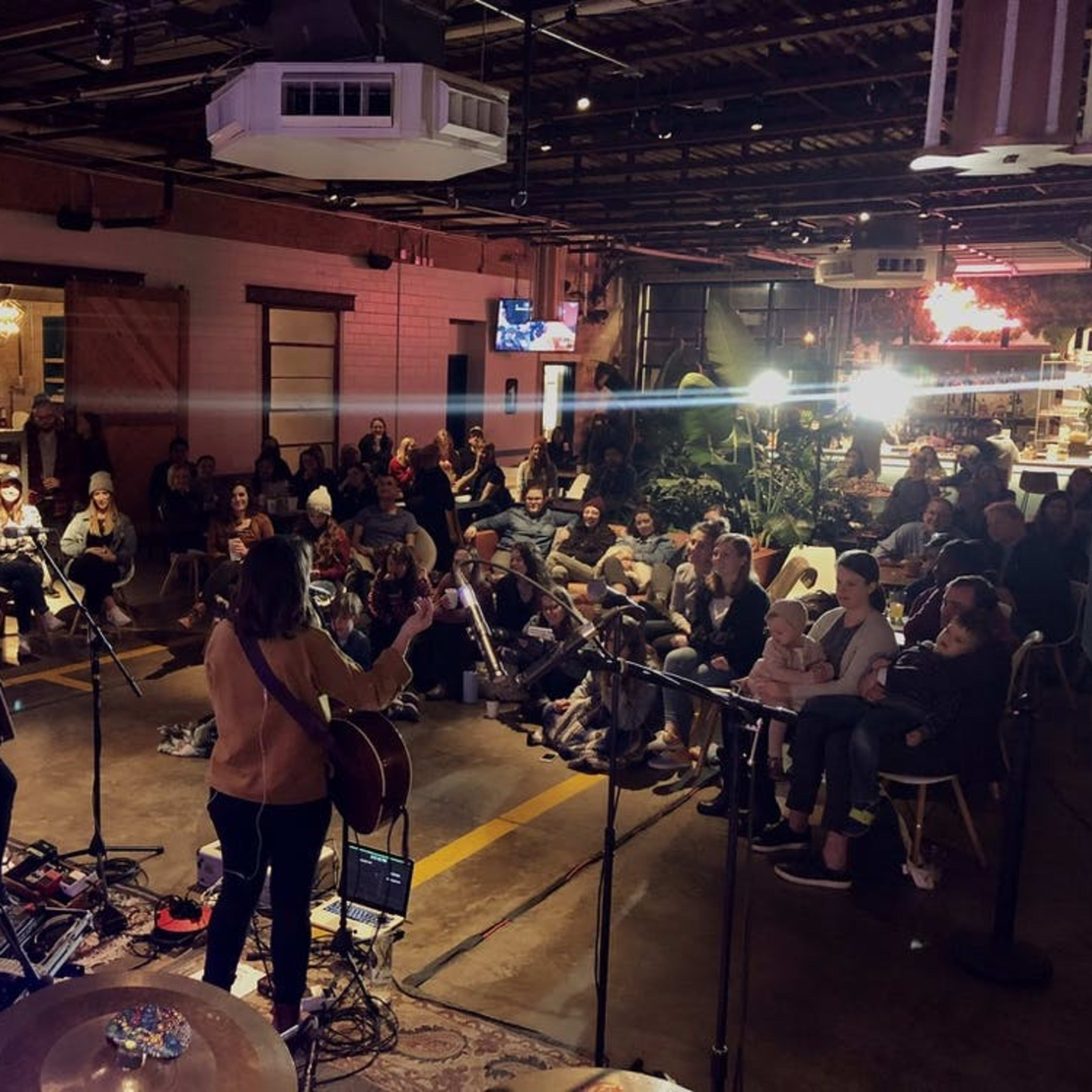 Two musicians perform on stage in a dimly lit venue while an audience sits facing them.