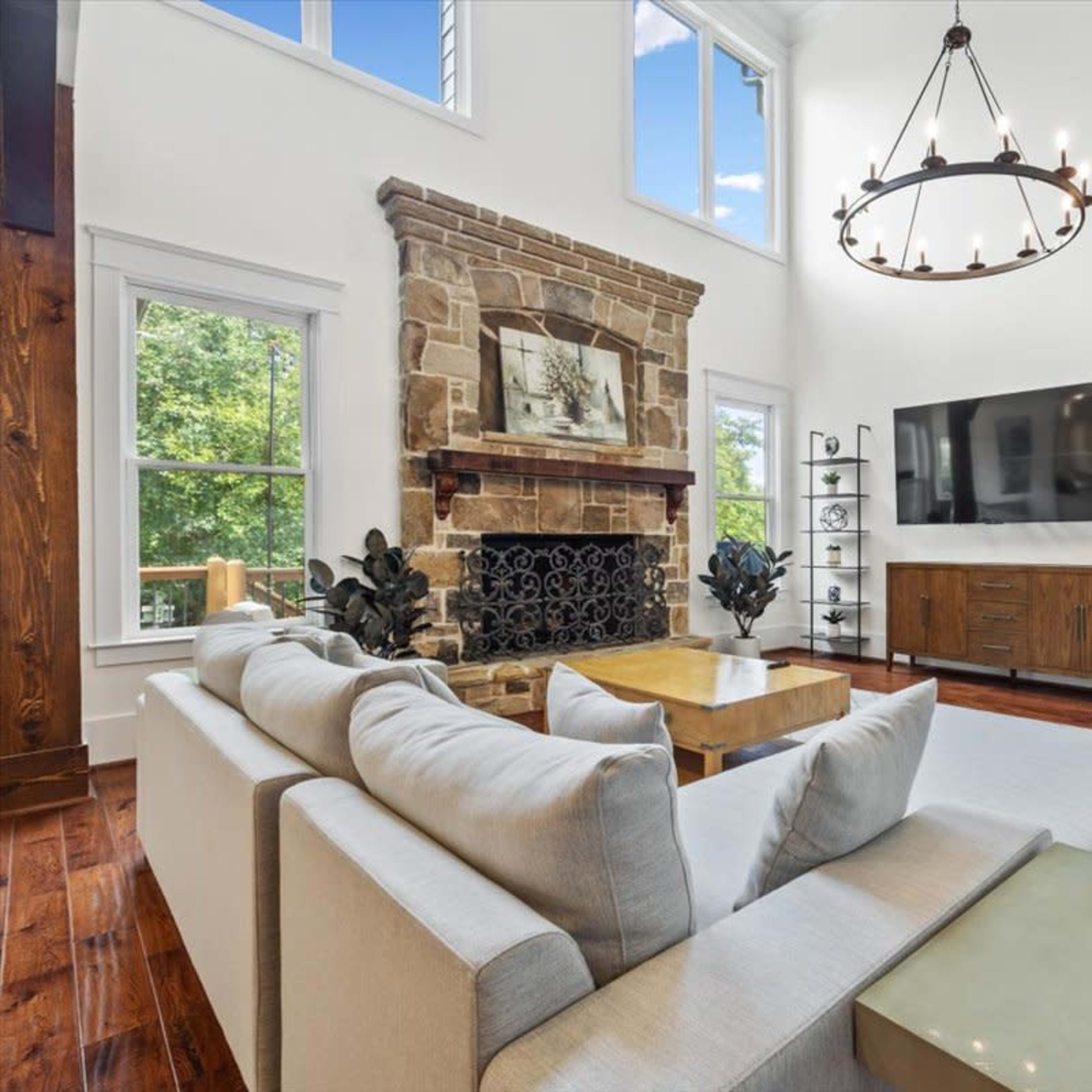 A spacious living room with a stone fireplace, large windows, and a modern TV setup on a wooden media console.
